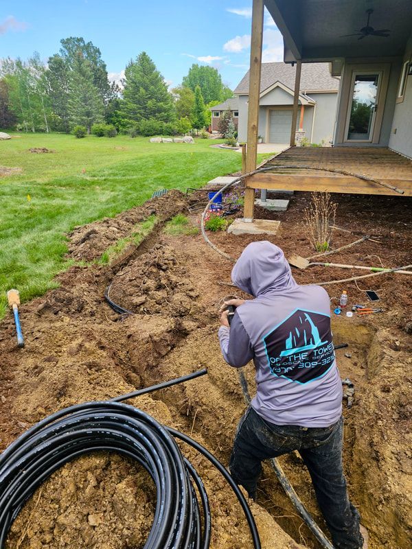 Person digging a trench in a backyard, near a deck. Black pipes and coil of cable visible.