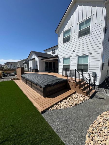 A backyard with a hot tub on a wooden deck next to a house with white siding and gravel pathway.