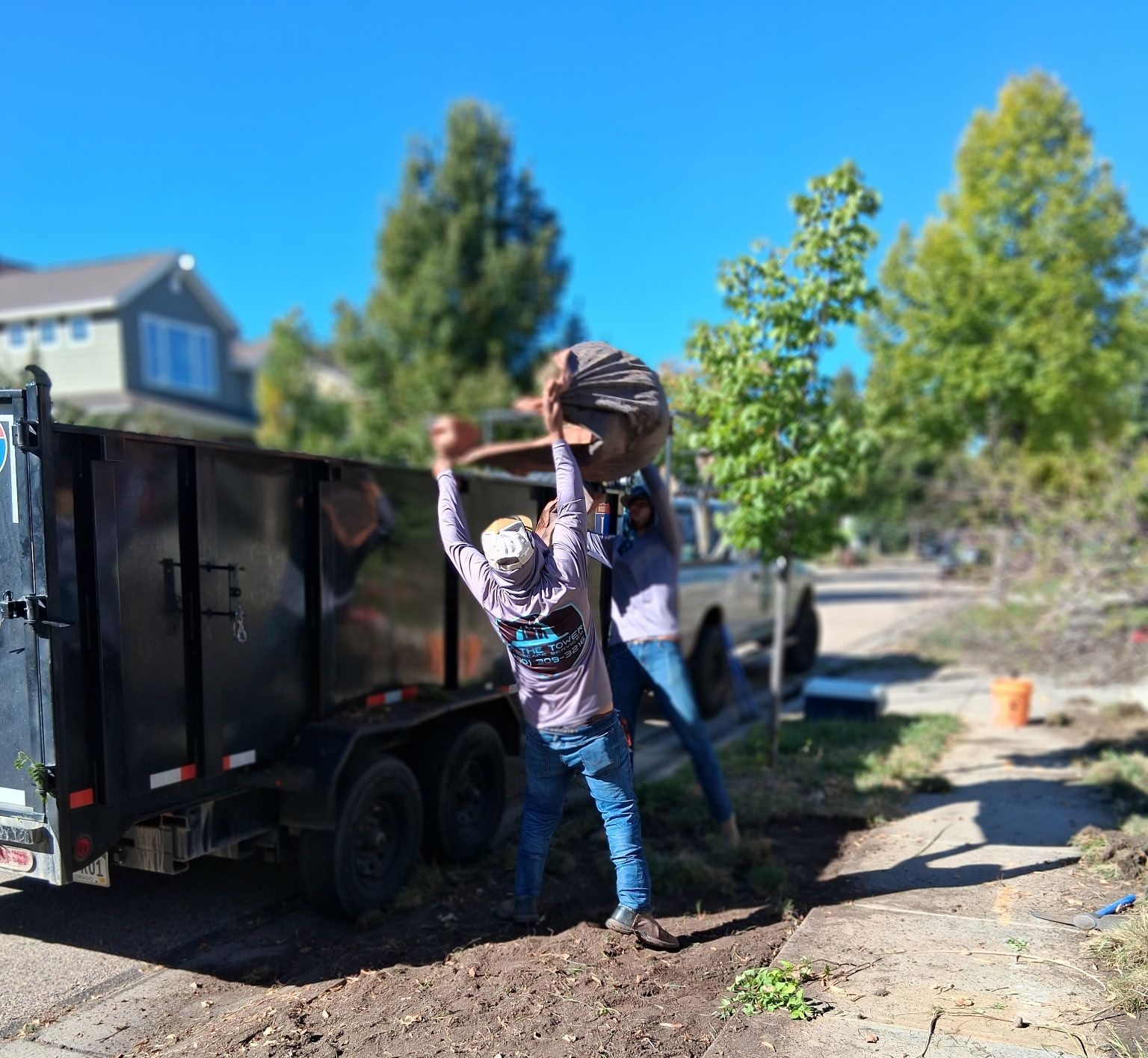 Two people loading a large trash bag into a black trailer on a sunny day.