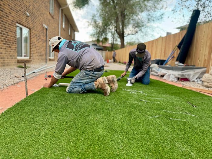 Two workers installing artificial turf in a yard, kneeling with tools. Green turf, brown shoes.