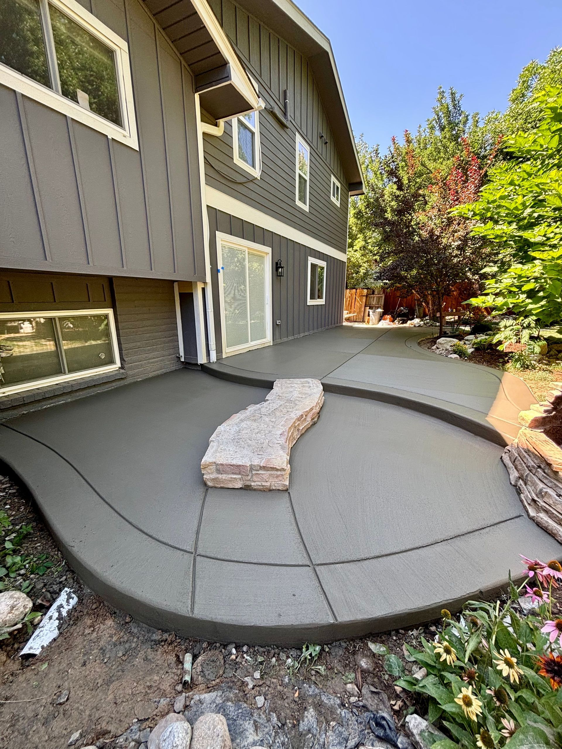 Newly poured concrete patio beside a two-story house, featuring a curved design and a large rock.