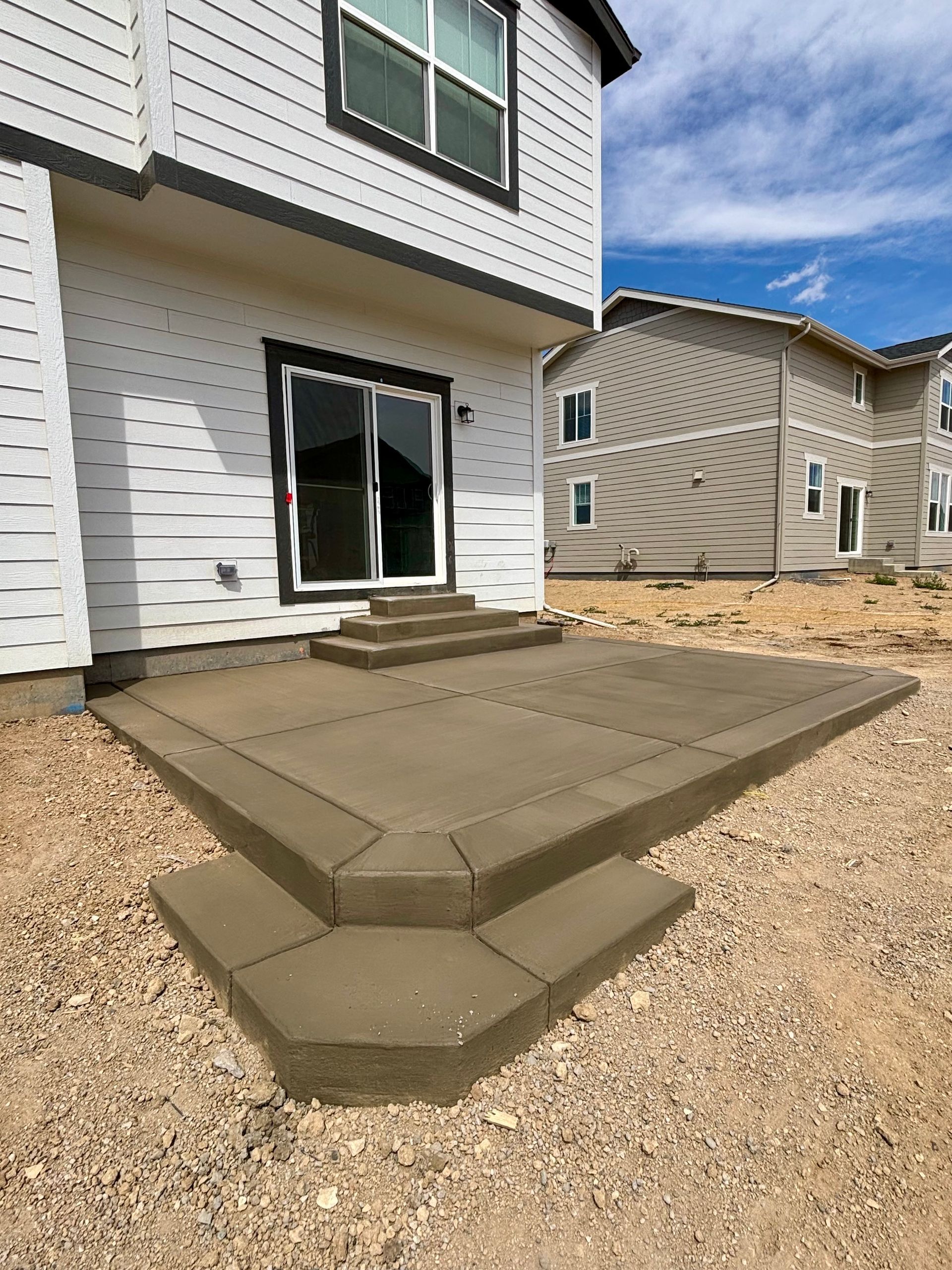 Concrete patio with steps, next to a two-story house with a sliding glass door and gravel ground.