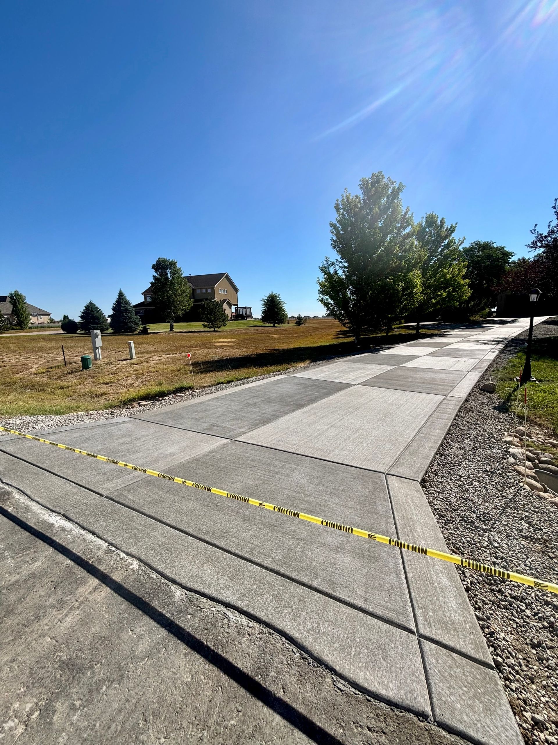 Concrete driveway under bright blue sky, yellow caution tape, vacant lots with houses in background.