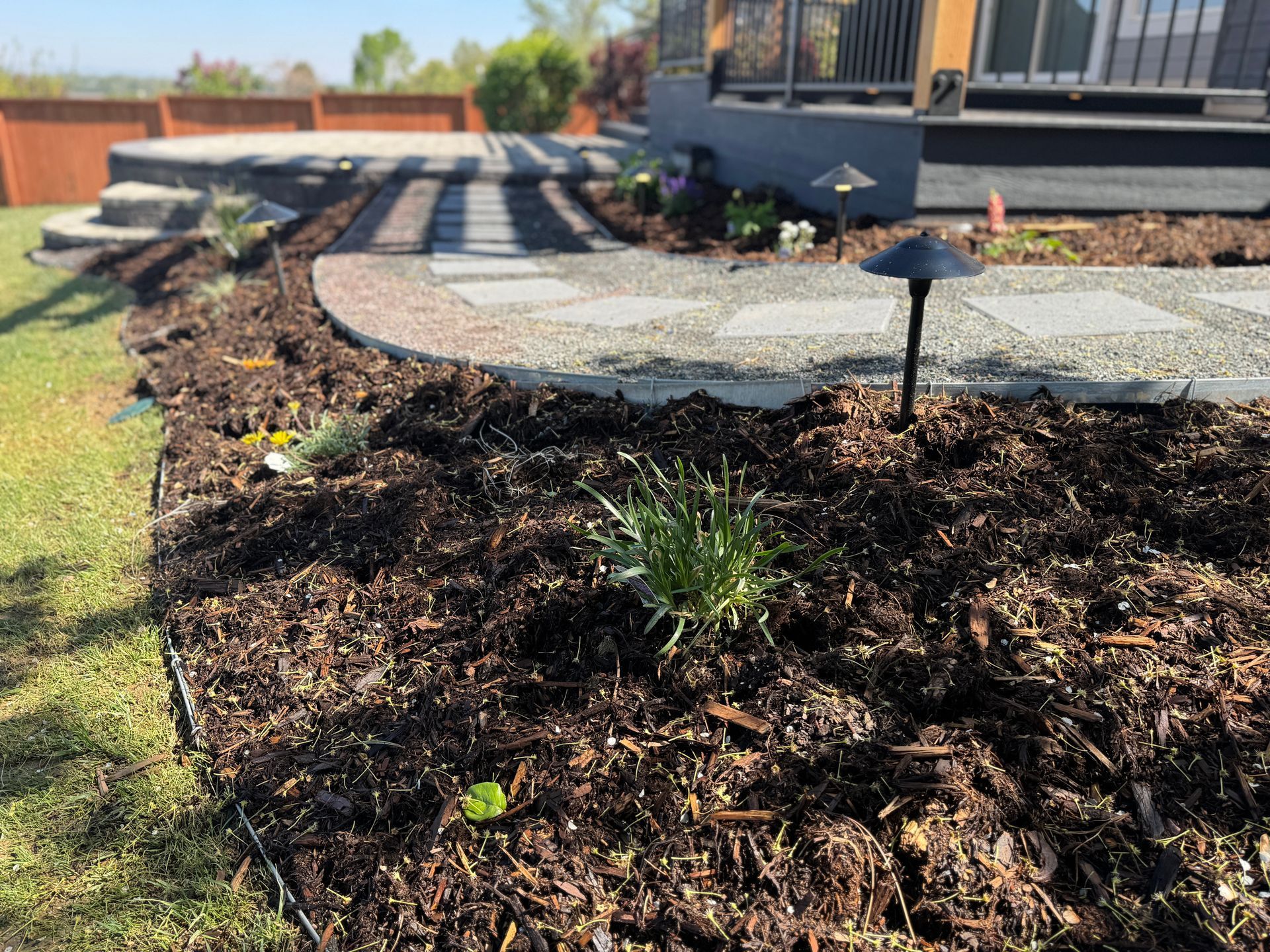 Garden bed with mulch, stepping stones leading to a patio, and a lawn in the background.