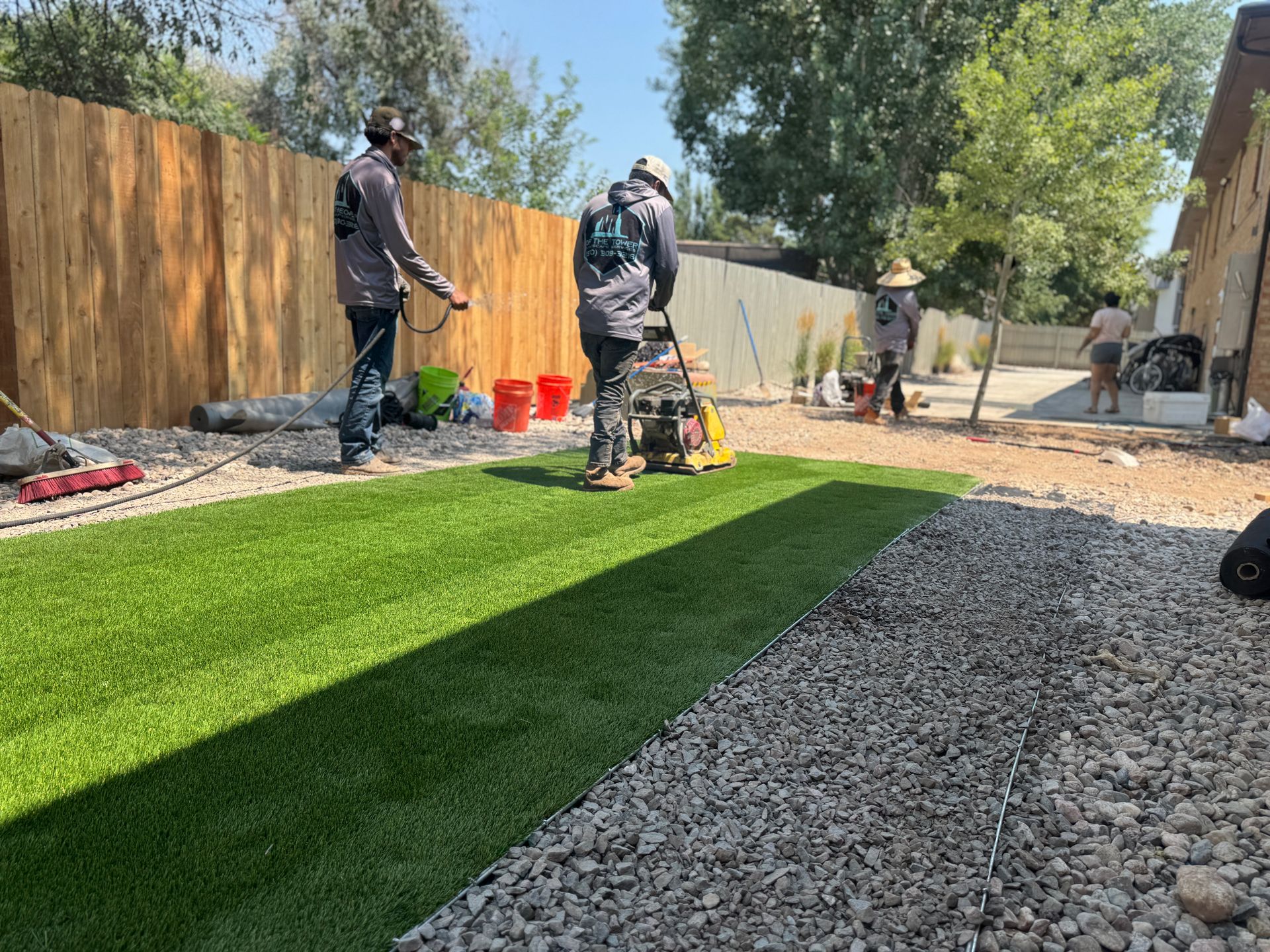 Workers install artificial turf in a backyard with a gravel border.