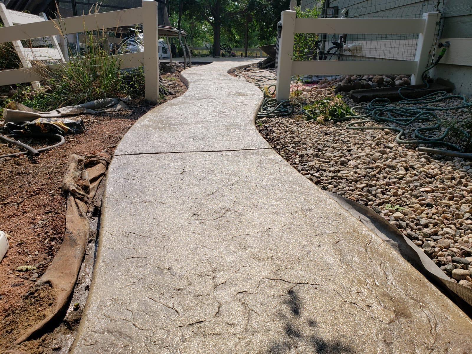 Concrete pathway through a yard, bordered by rocks and a white fence.