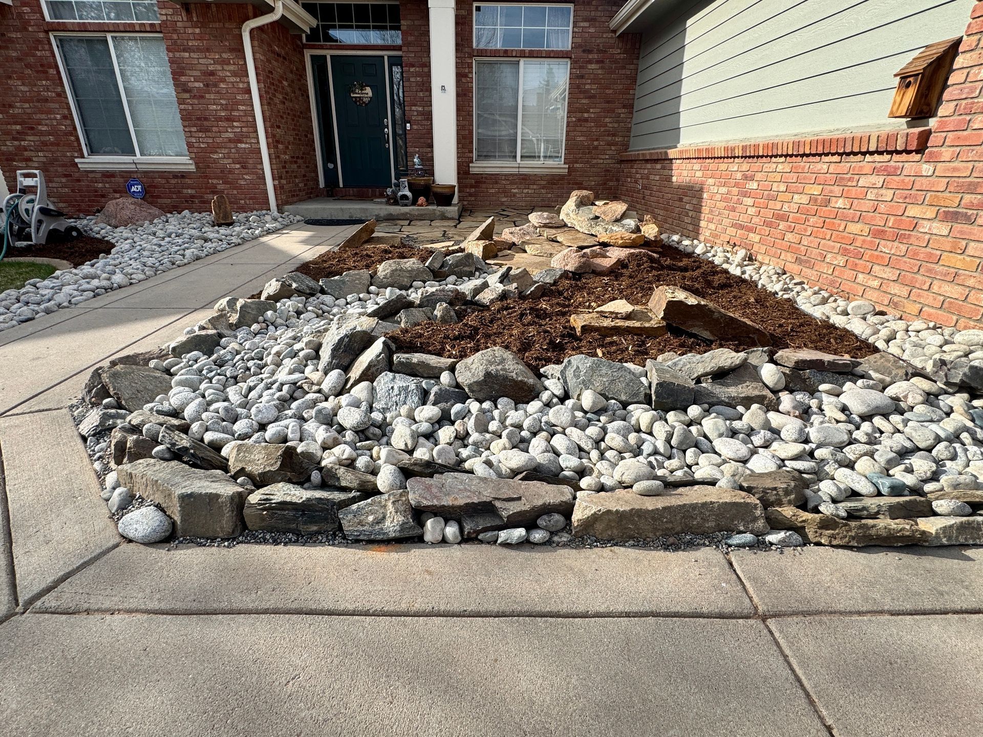 A front yard rock garden with a brick house. Grey rocks surround a mulched planting area.
