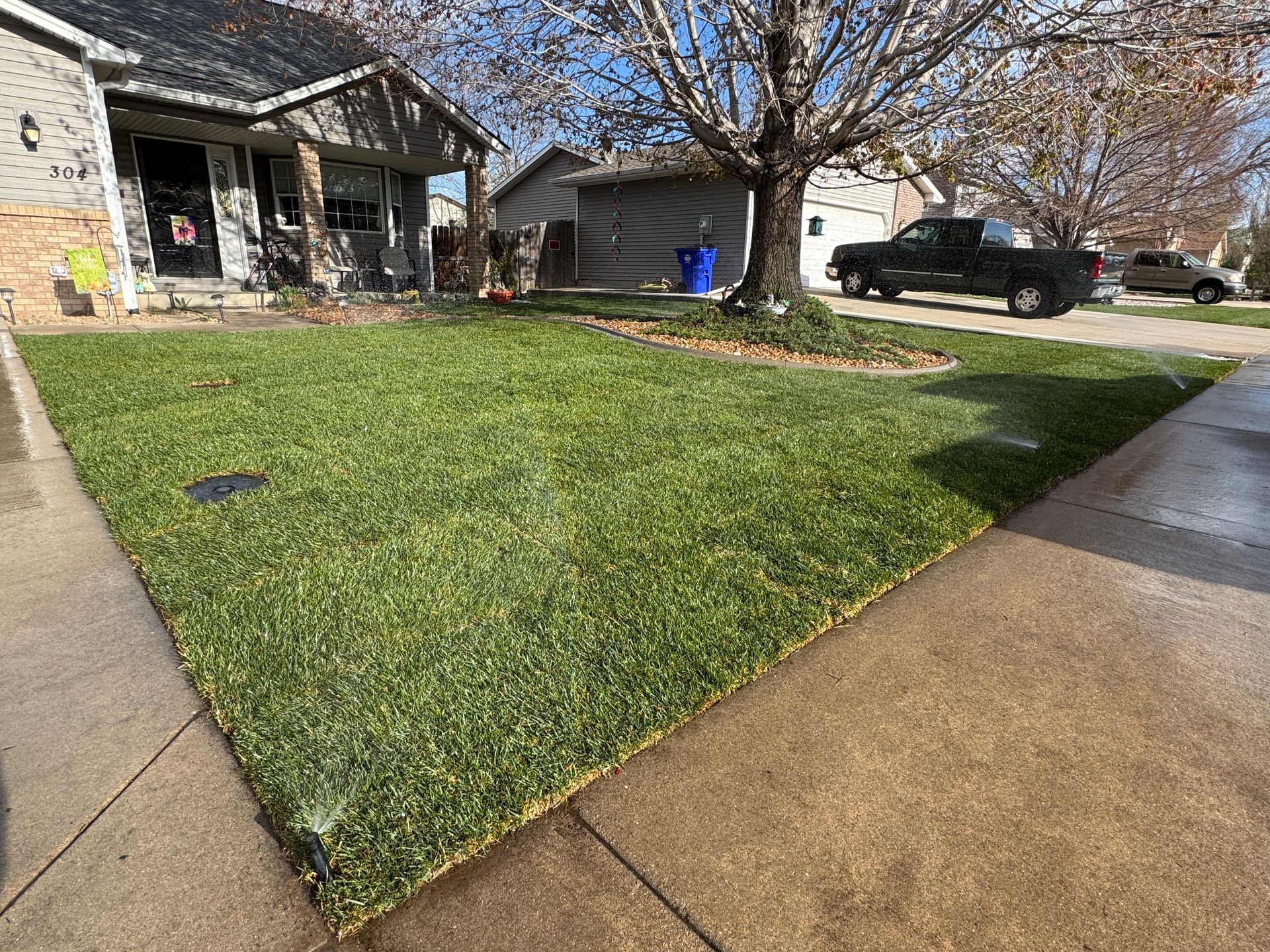 A sprinkler waters a green lawn in front of a house, with a truck and cars parked nearby.