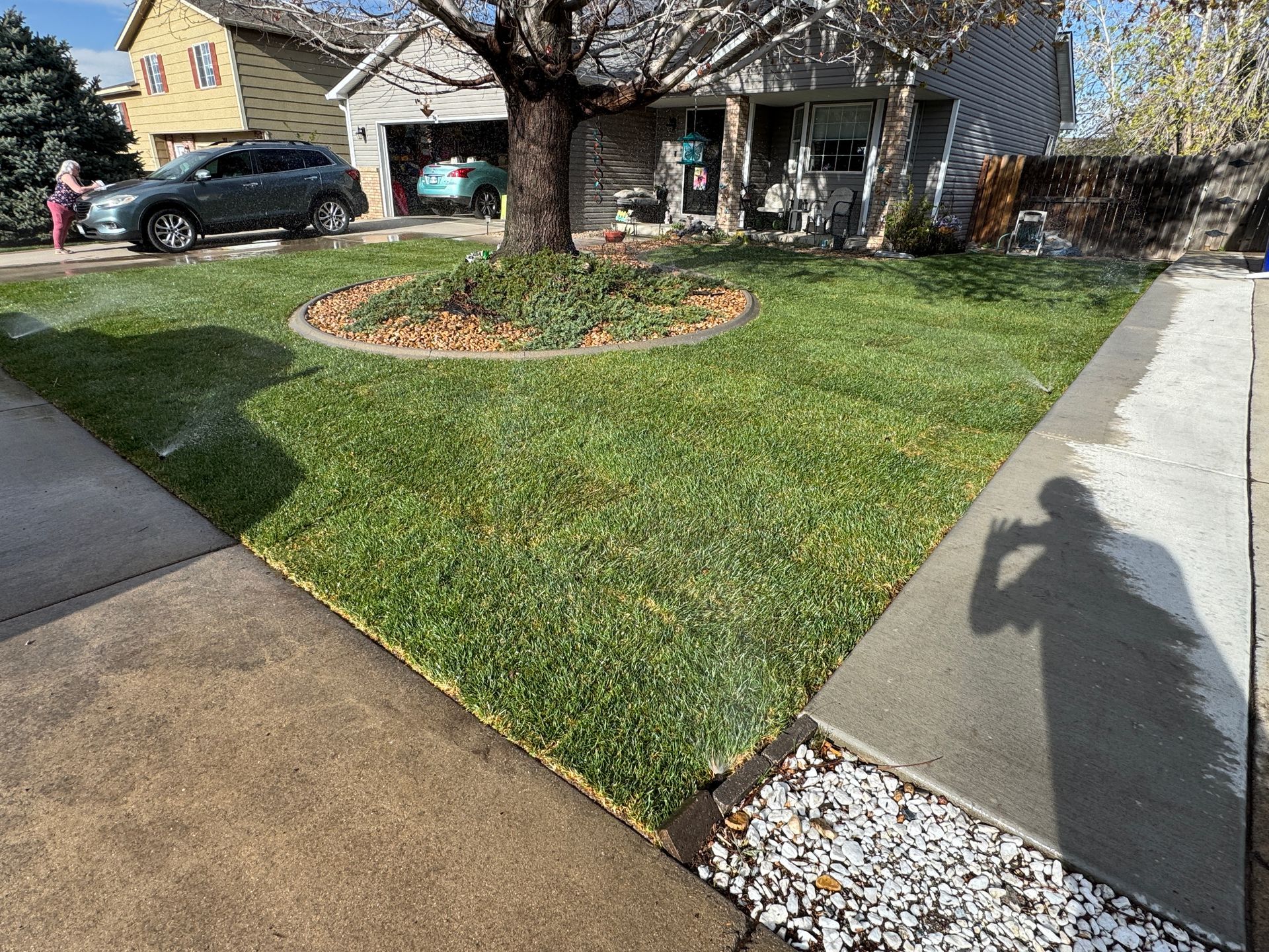 Green lawn being watered by sprinklers in front of a house, with a parked car and person visible.