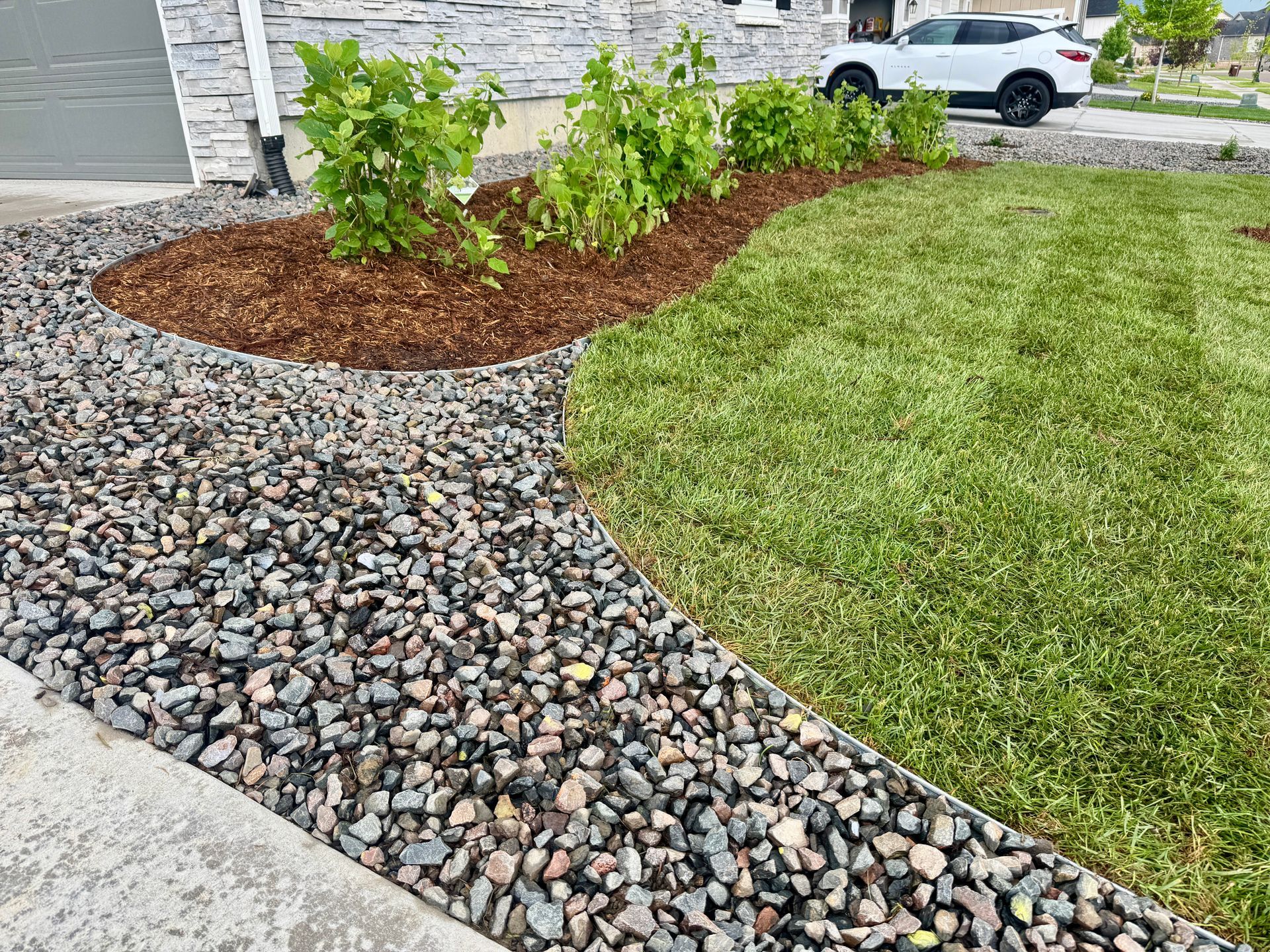 A landscaped yard with a gravel path, grass, and plants bordered by wood chips. A car is parked nearby.