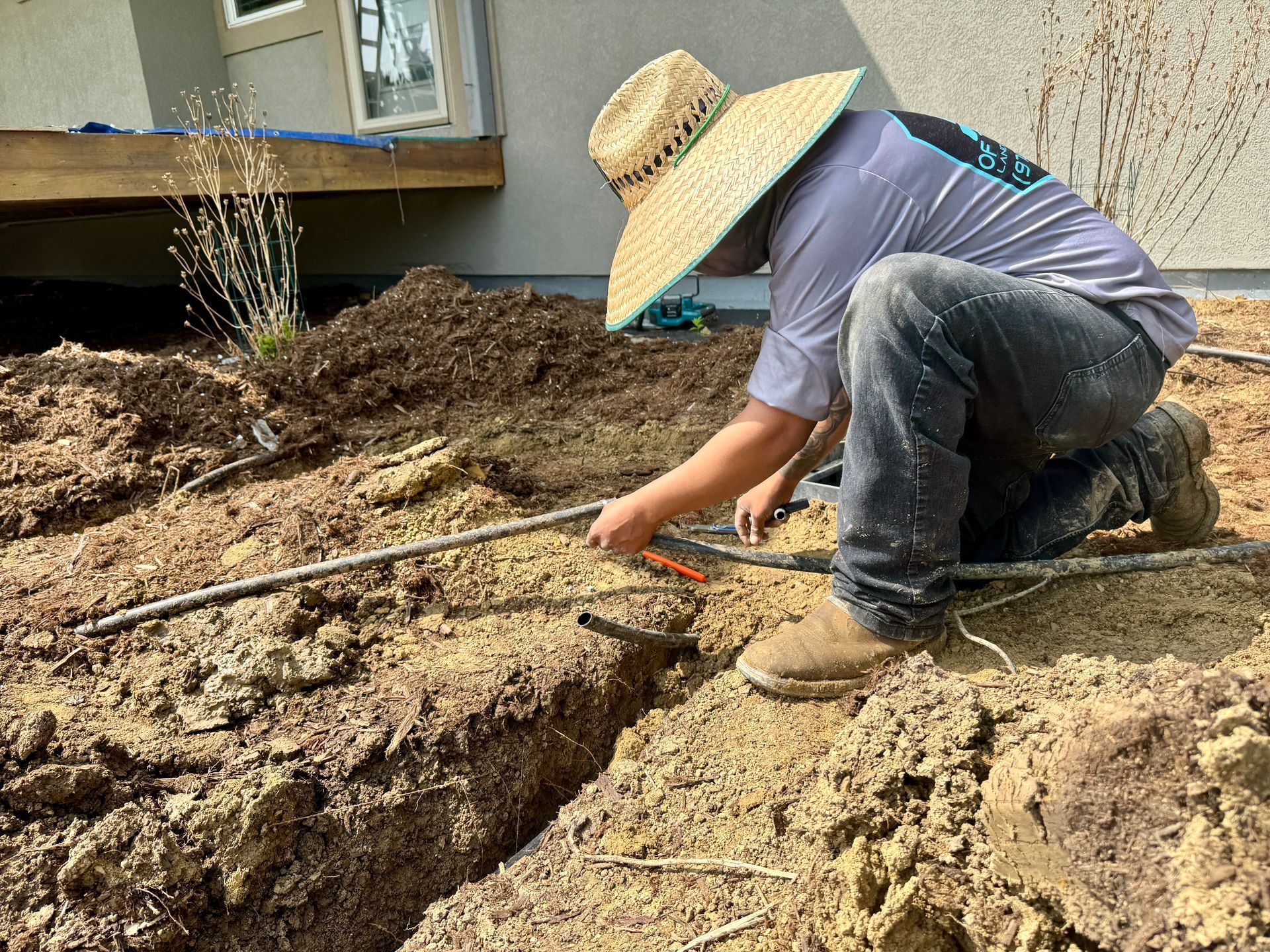 Man in straw hat working on irrigation system trench in front of a house.