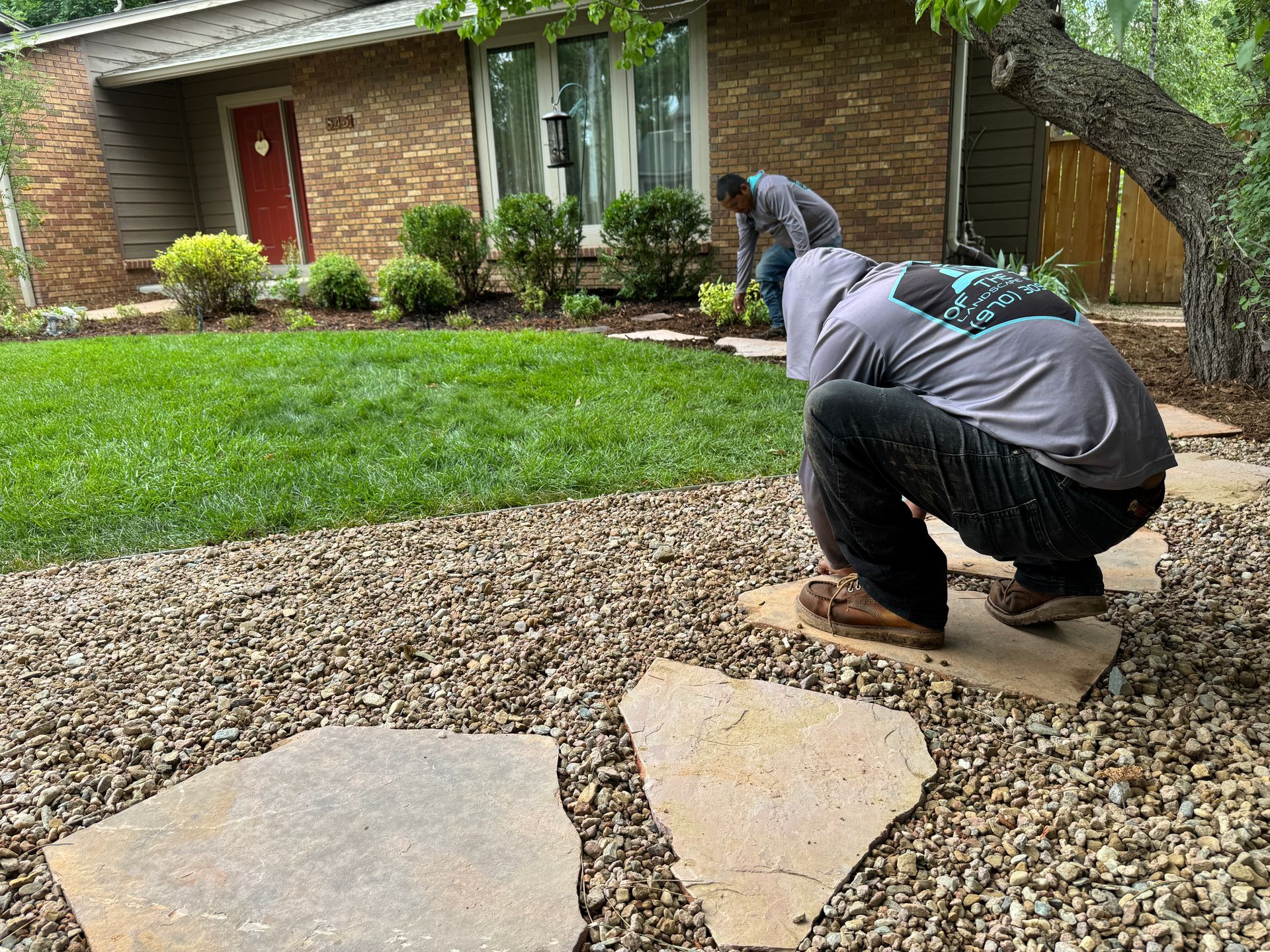 Two people working on landscaping a home; stepping stones, gravel, grass, and bushes visible.