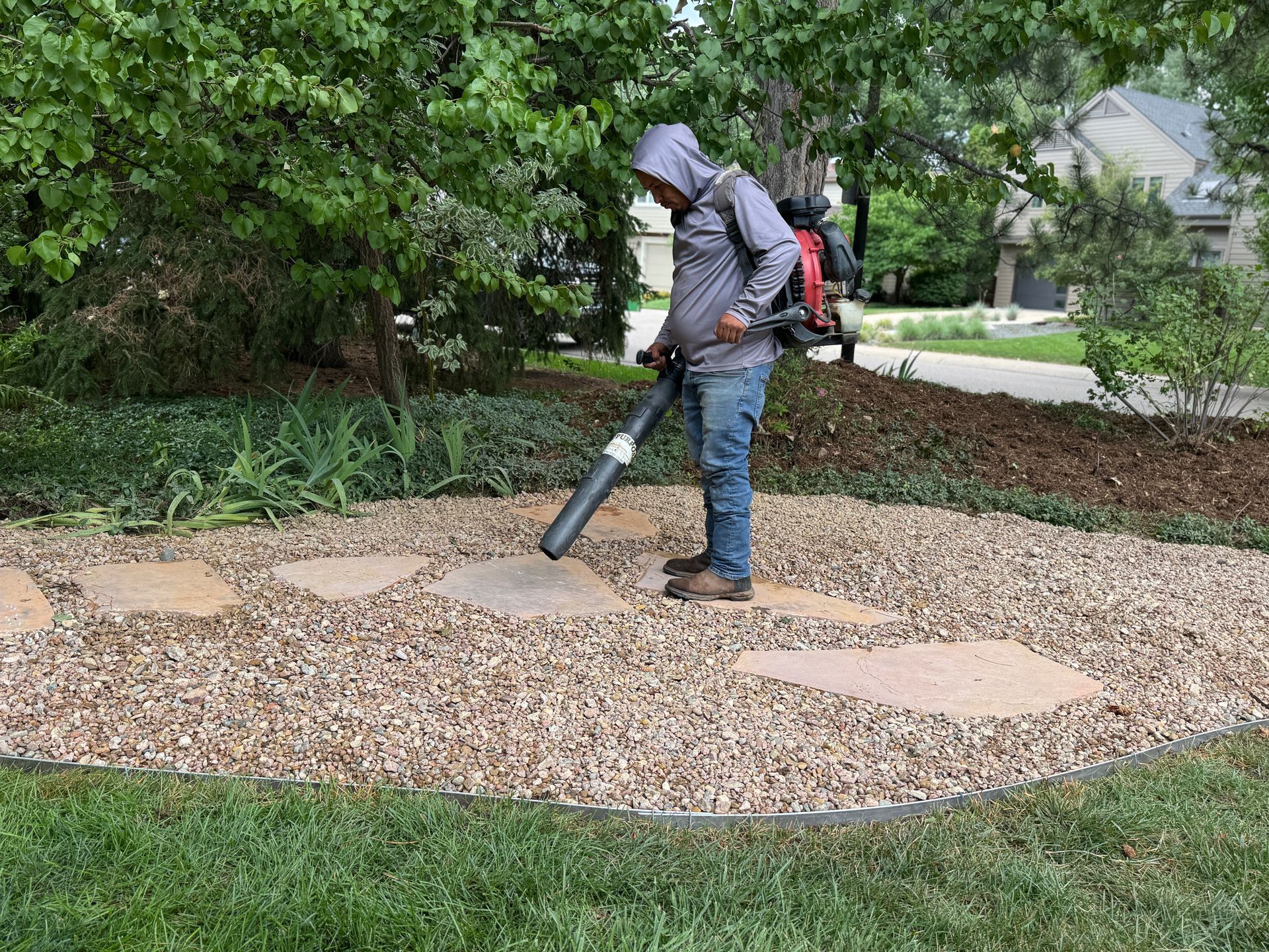 Man using a backpack leaf blower on a gravel path with stepping stones in a landscaped yard.