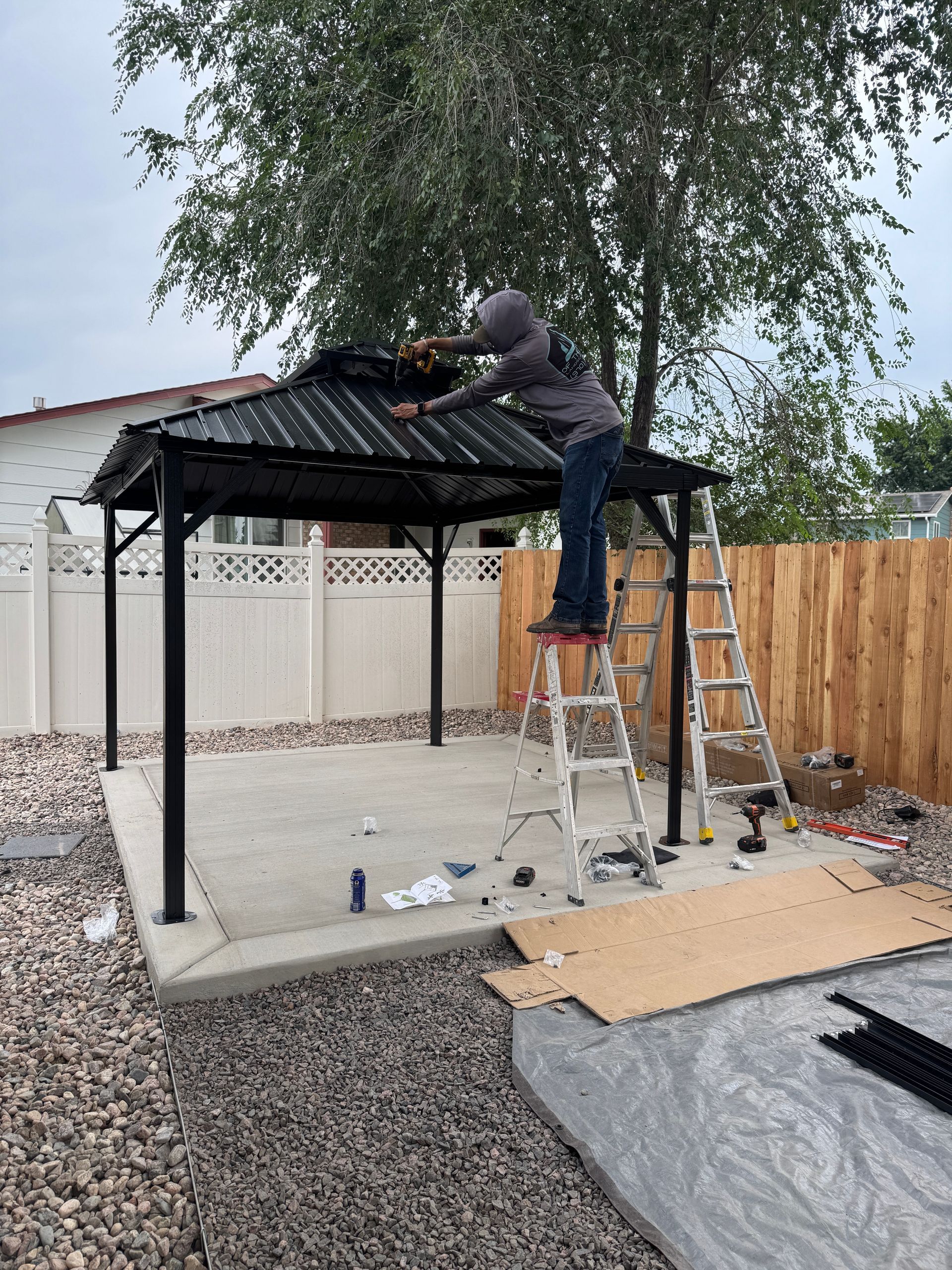 Person on a ladder assembles a black gazebo on a concrete patio in a backyard setting.