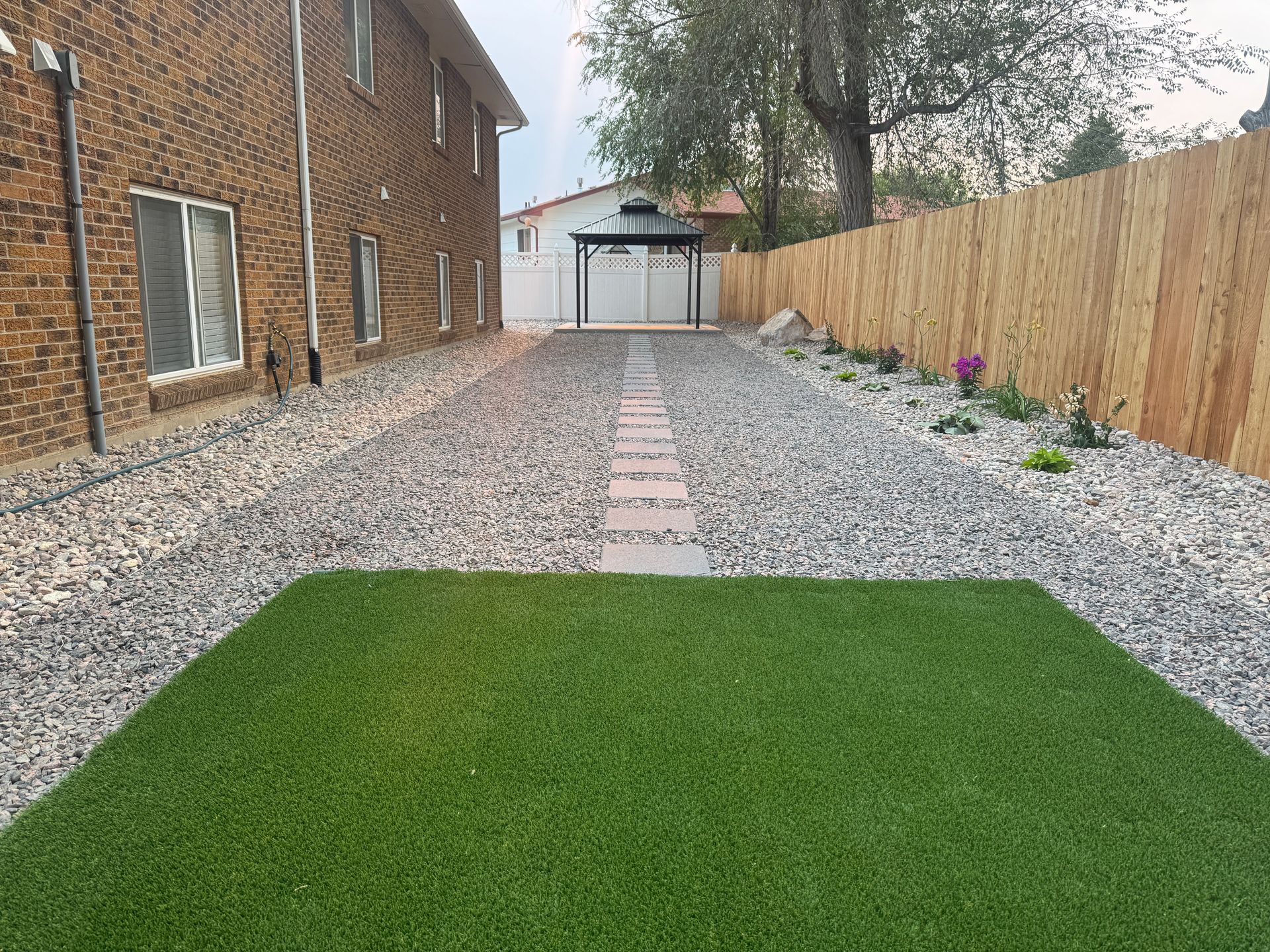 Driveway with gravel, a path, a lawn patch, and a brick building next to a wooden fence.