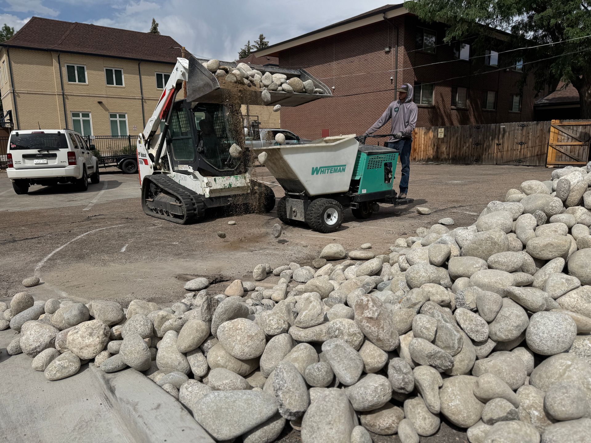 Bobcat loader dumping rocks into a motorized cart, man guiding. Rocks and buildings in the background.