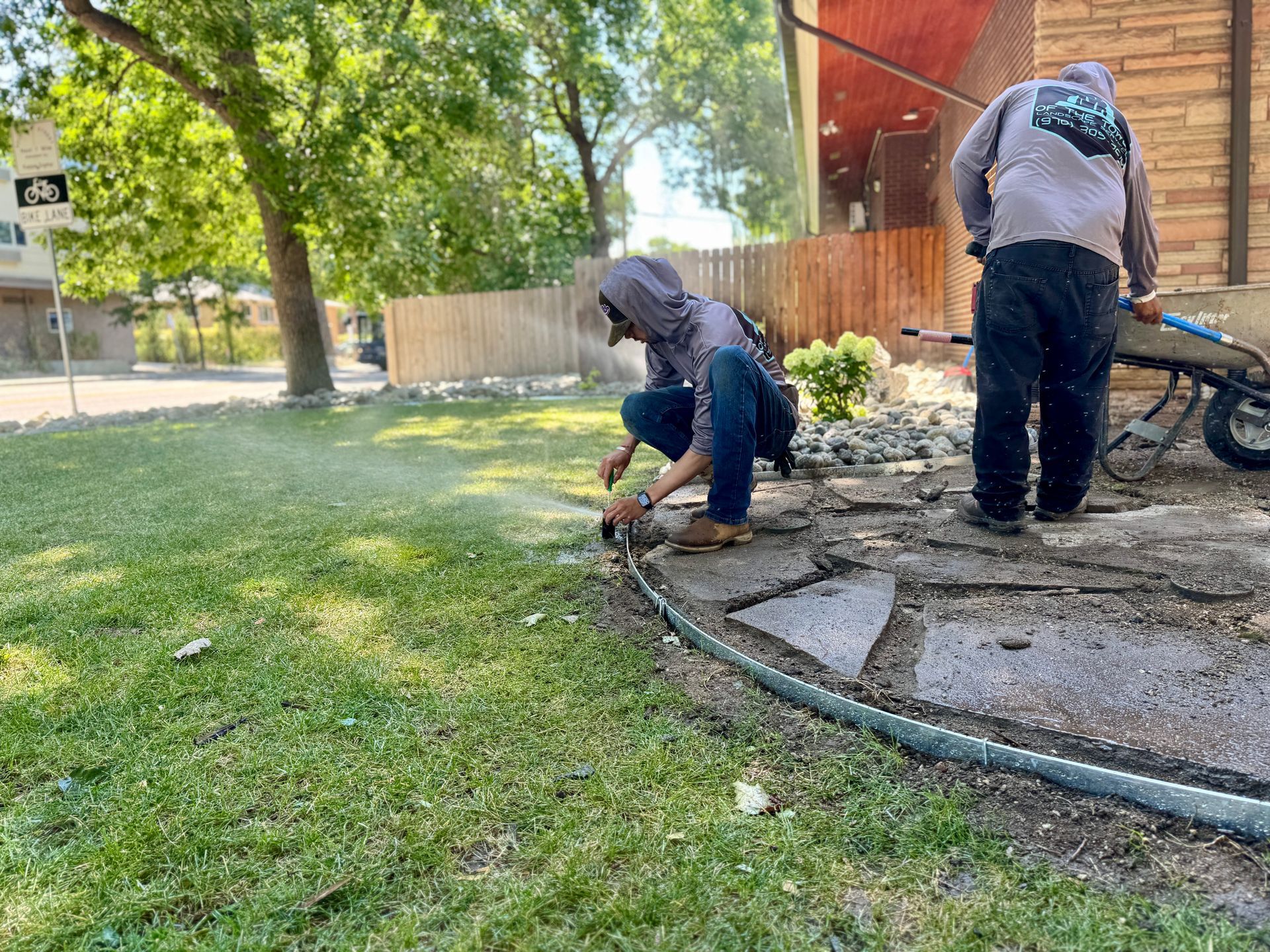 Two people installing sprinklers in a yard; one crouches near hose, other stands near a wheelbarrow.