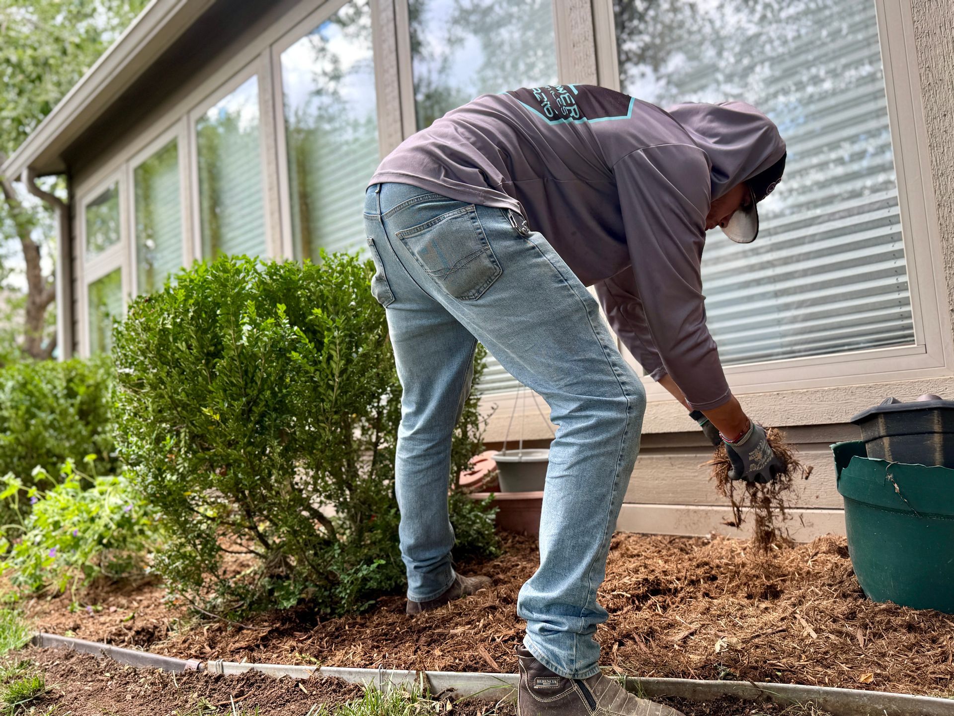A person in a hoodie and jeans mulches the garden bed next to a house.