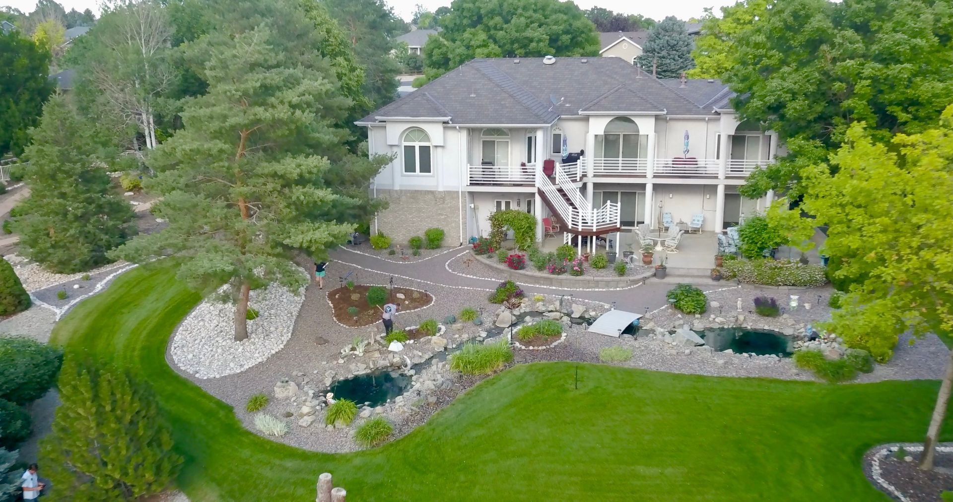 White house with multiple levels, balcony, and staircase, with lush landscaping and ponds.