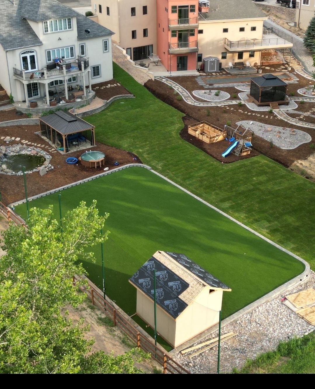 Aerial view of a terraced yard with a green artificial lawn, play area, pond, and various buildings.