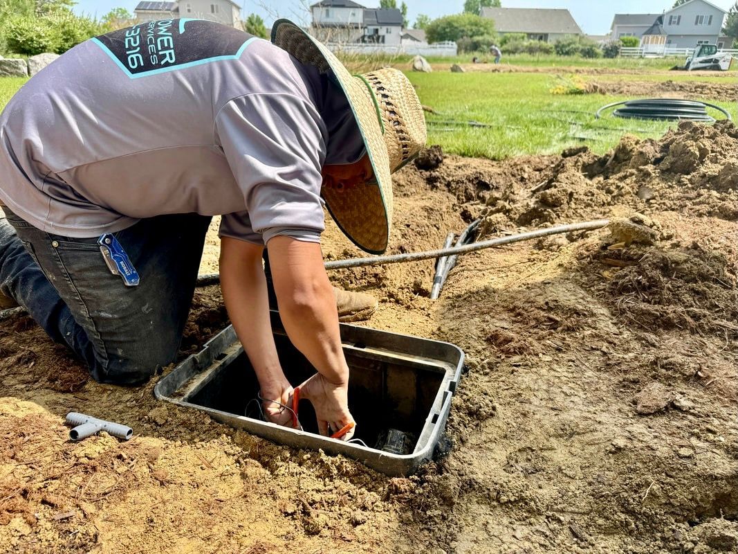 Person working on sprinkler system in a brown, muddy yard wearing a straw hat and gray shirt.