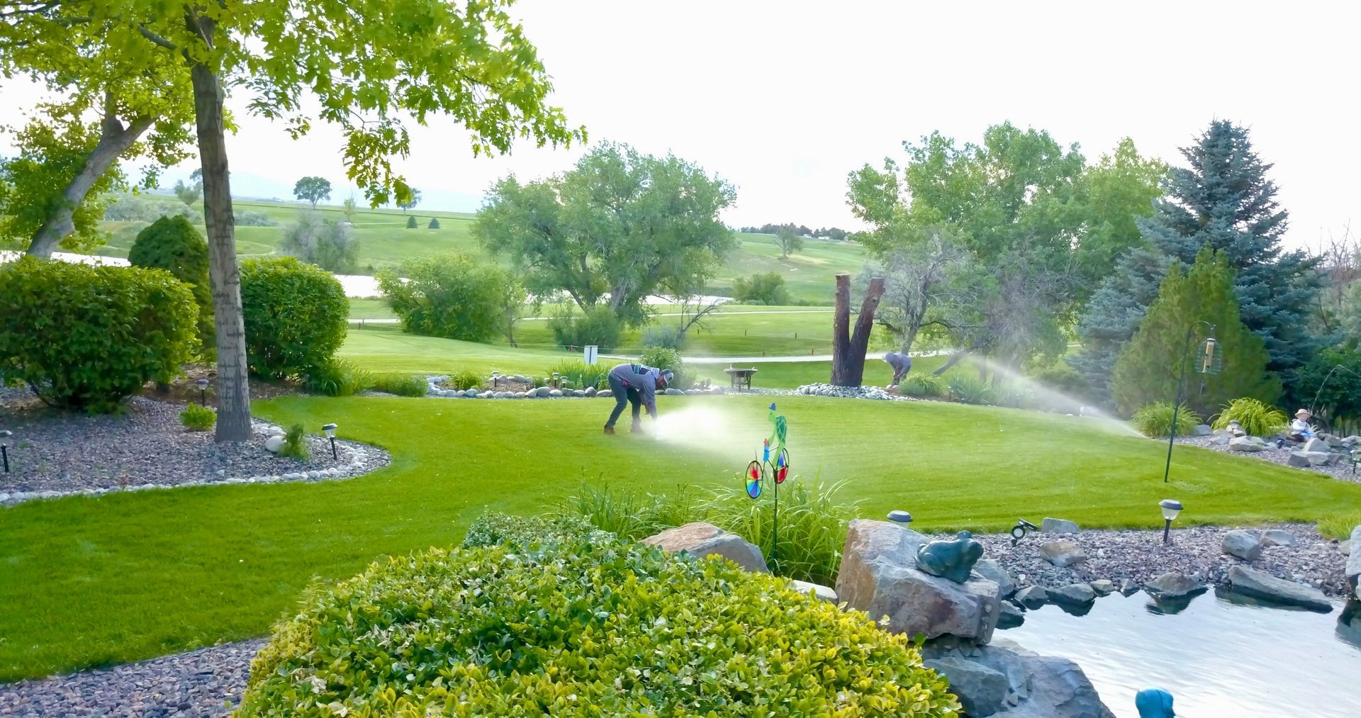 Man tending to a sprinkler on a green lawn, surrounded by trees and a pond in a landscaped yard.