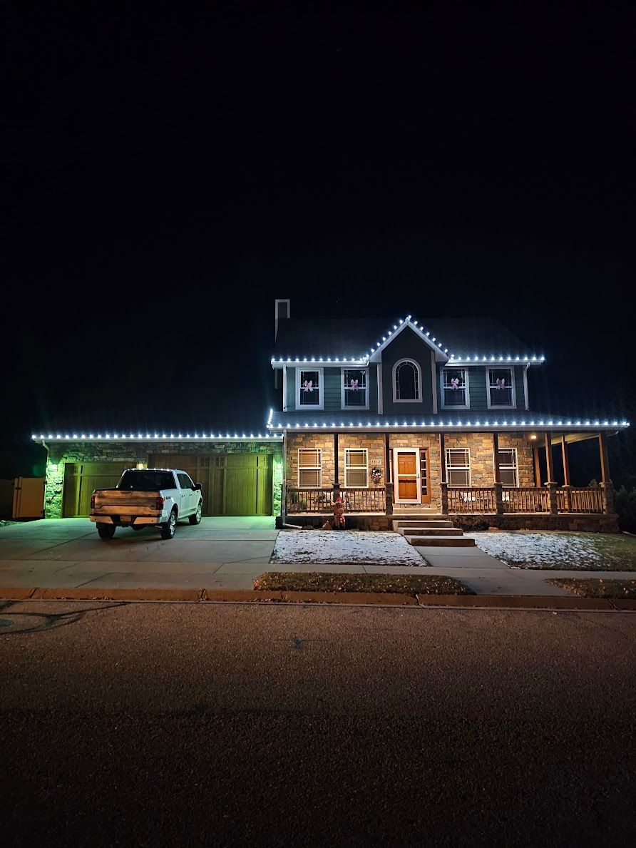 A two-story house at night with white lights along the roof and porch, a truck in the garage.