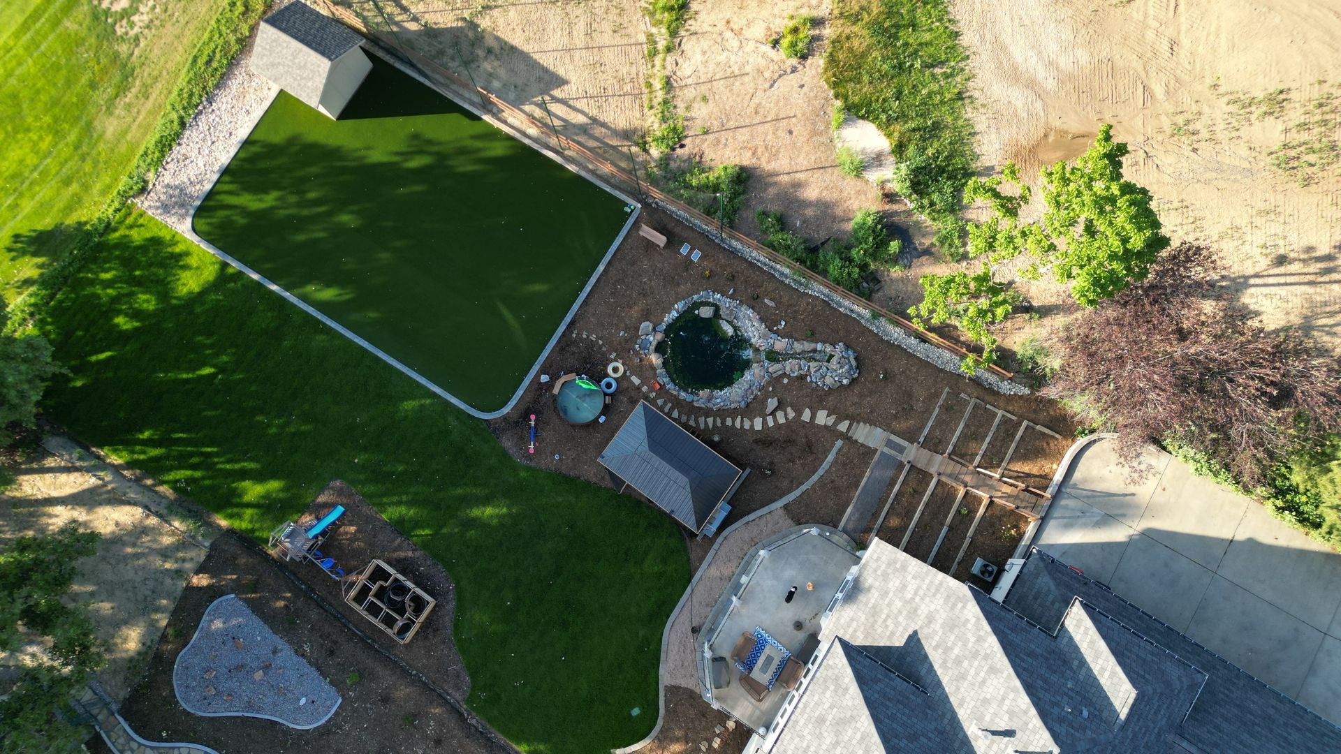 Overhead view of a backyard with a pool, garden, and house. Green grass, brown mulch, and blue water are visible.