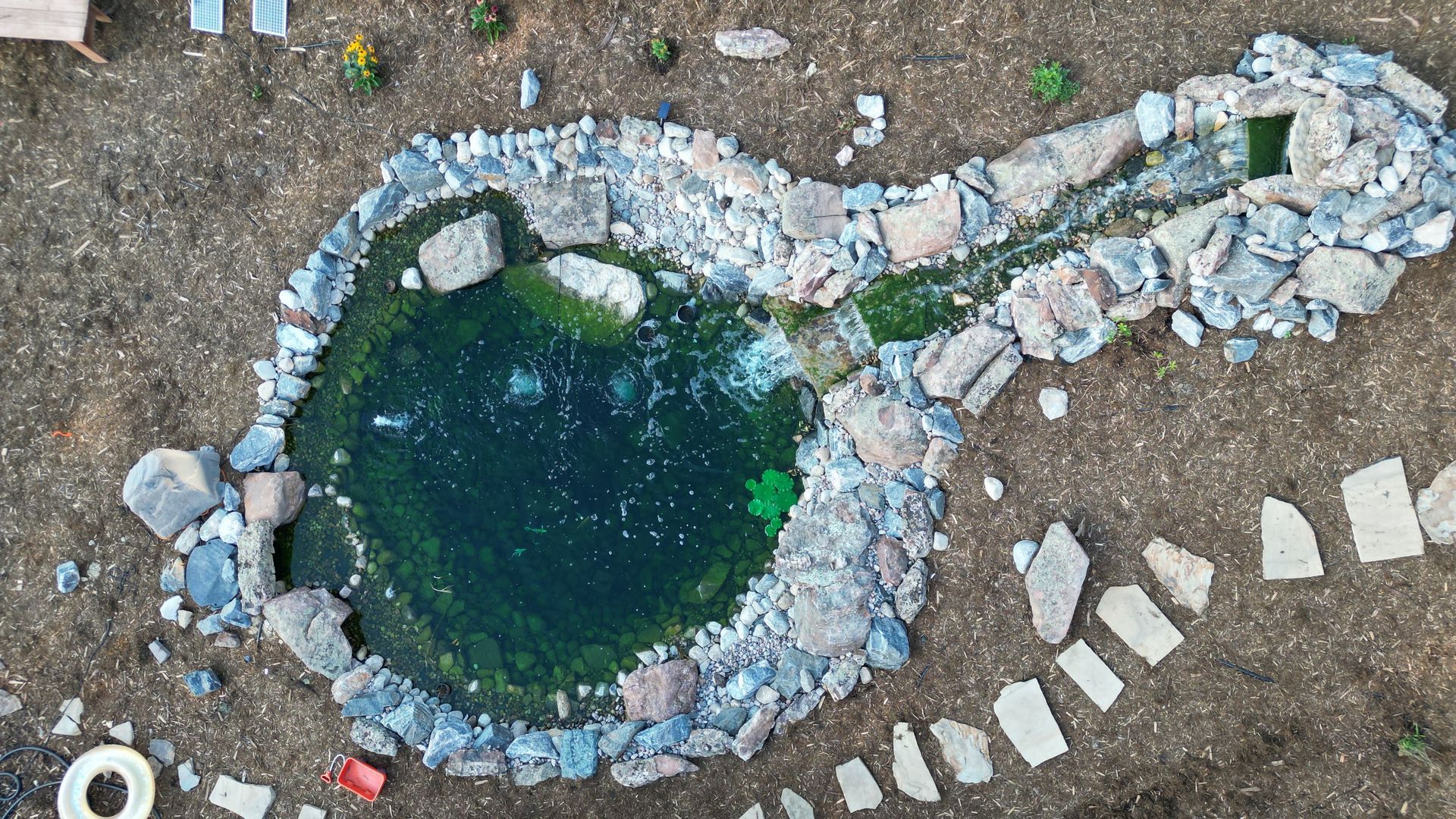 Overhead view of a rock-lined pond with dark green water, and a small waterfall.