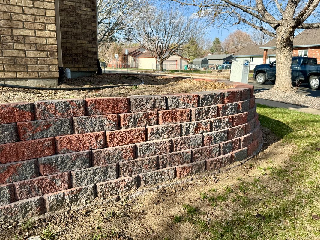 A curved, multi-toned brick retaining wall in front of a building, grass in foreground, trees and houses in the background.