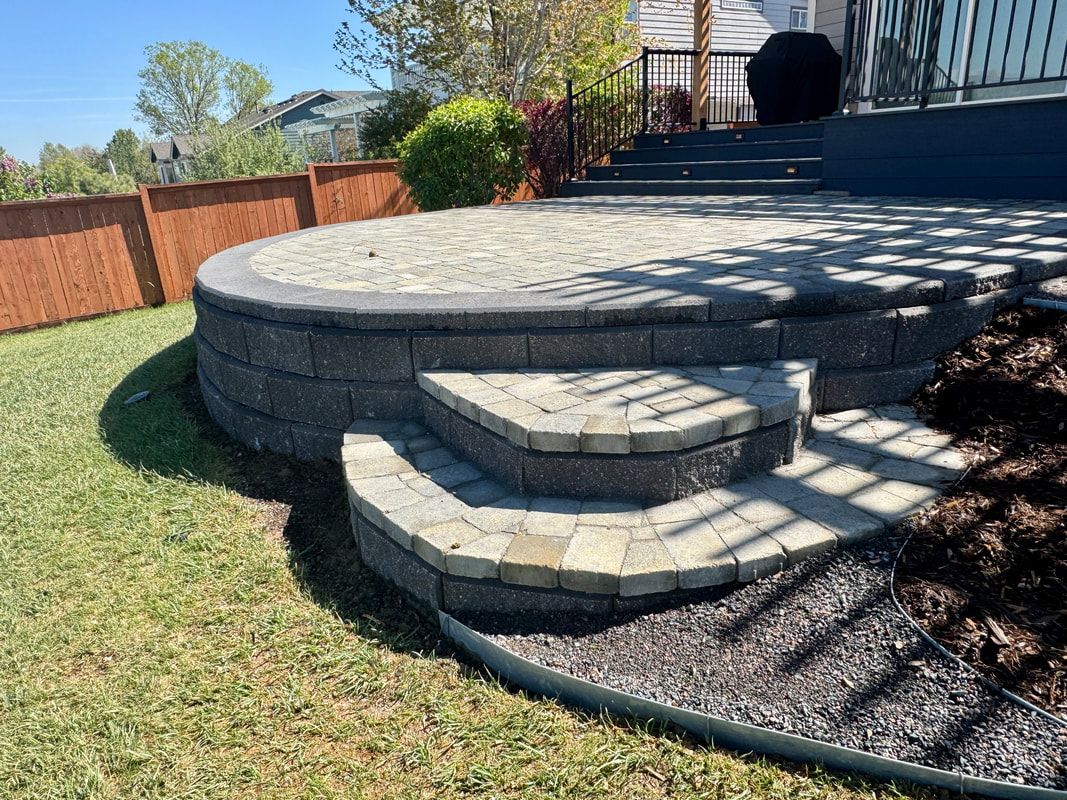Stone patio with steps, bordered by a retaining wall, and leading to a deck; grassy yard.