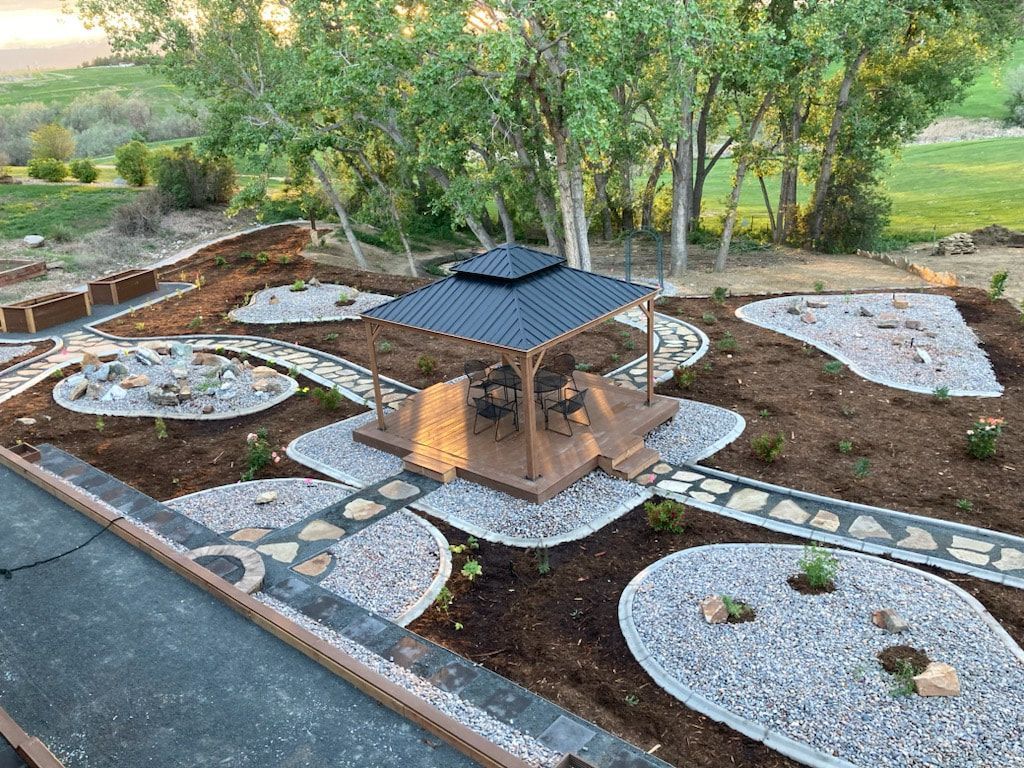 Gazebo with dark roof, surrounded by rock pathways and gardens in a backyard setting.