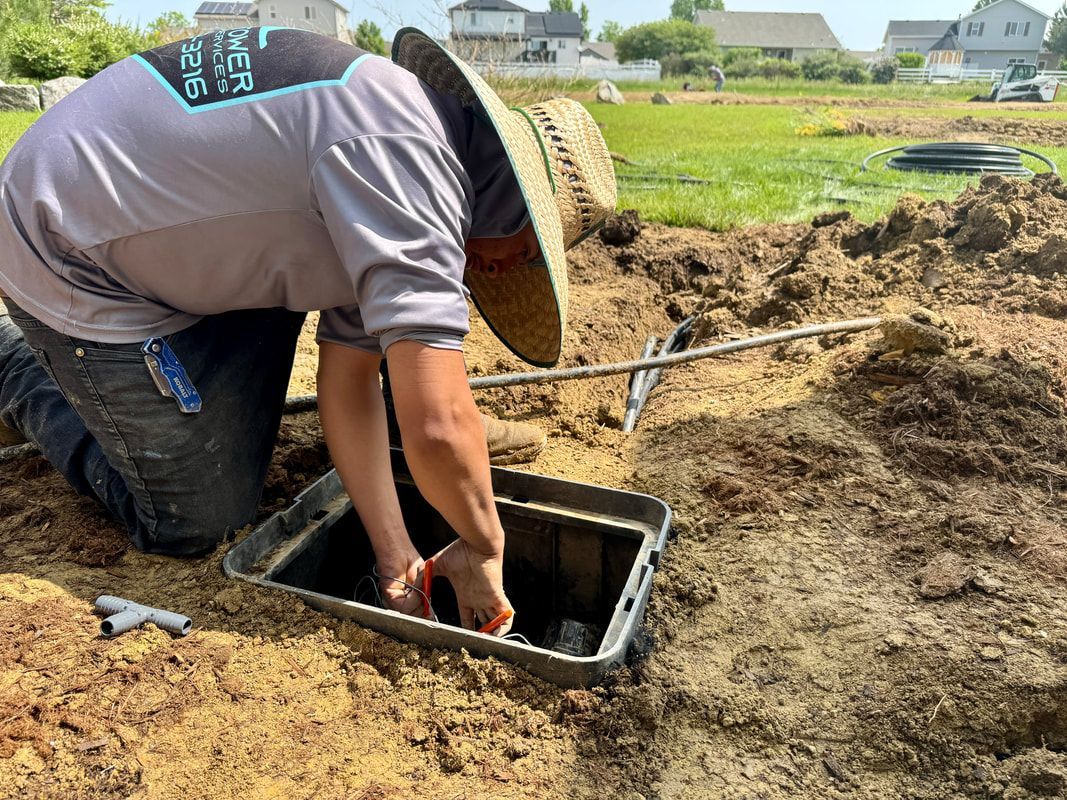 Person kneeling, working inside an irrigation control box in a yard, wearing a straw hat and gray shirt.