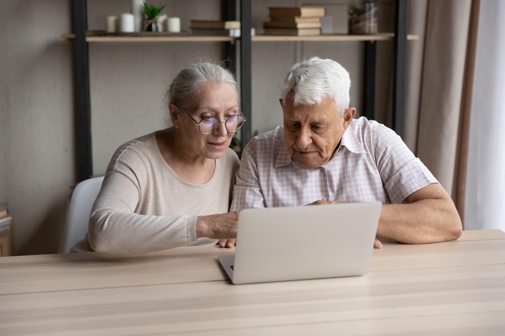 Two older adults sit at a table using a laptop in a bright living room.