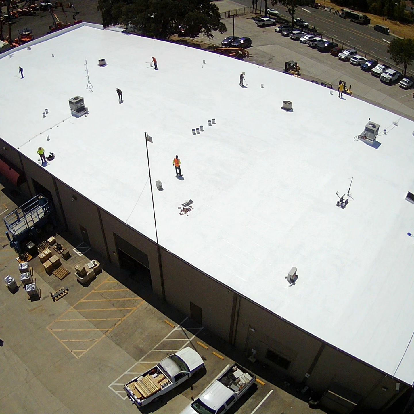 Aerial view of a large white-roofed building with rooftop equipment and parked cars nearby.
