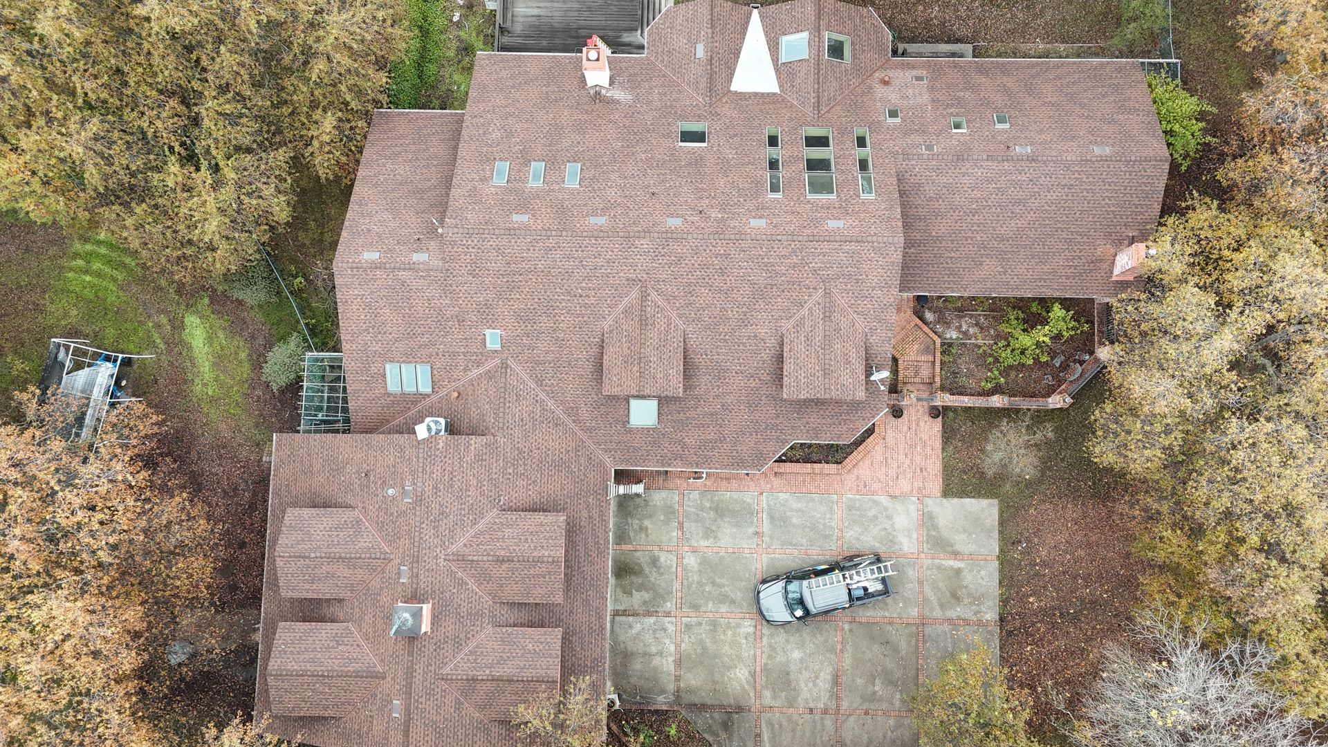 Aerial view of a large brown-roofed house with a car parked in the driveway, surrounded by trees.