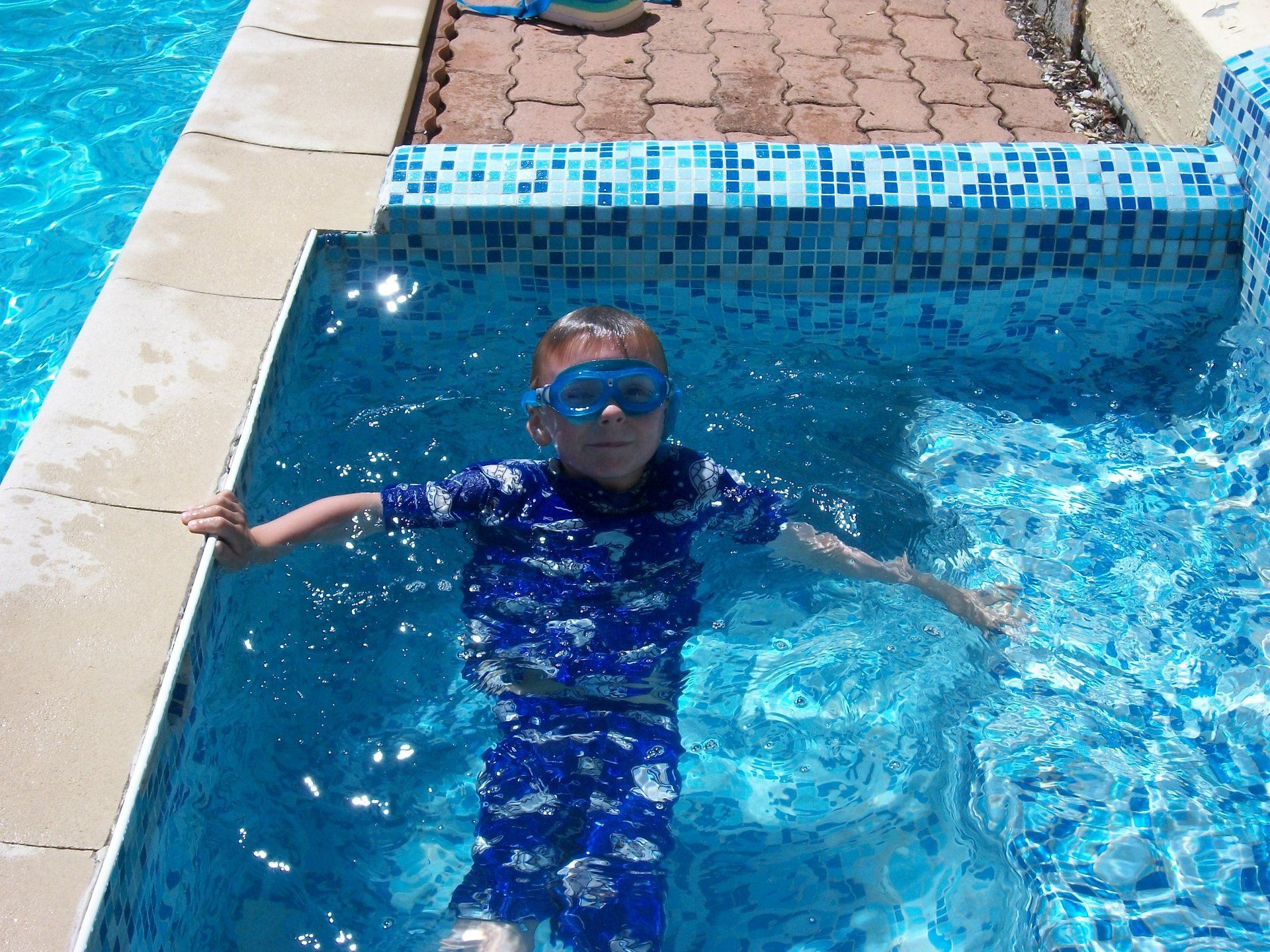 young boy wearing blue swim goggles and blue sun suit relaxing in a small tiled pool