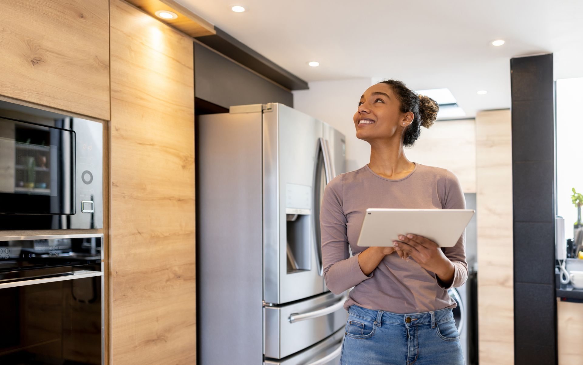 A woman is standing in a kitchen holding a tablet.