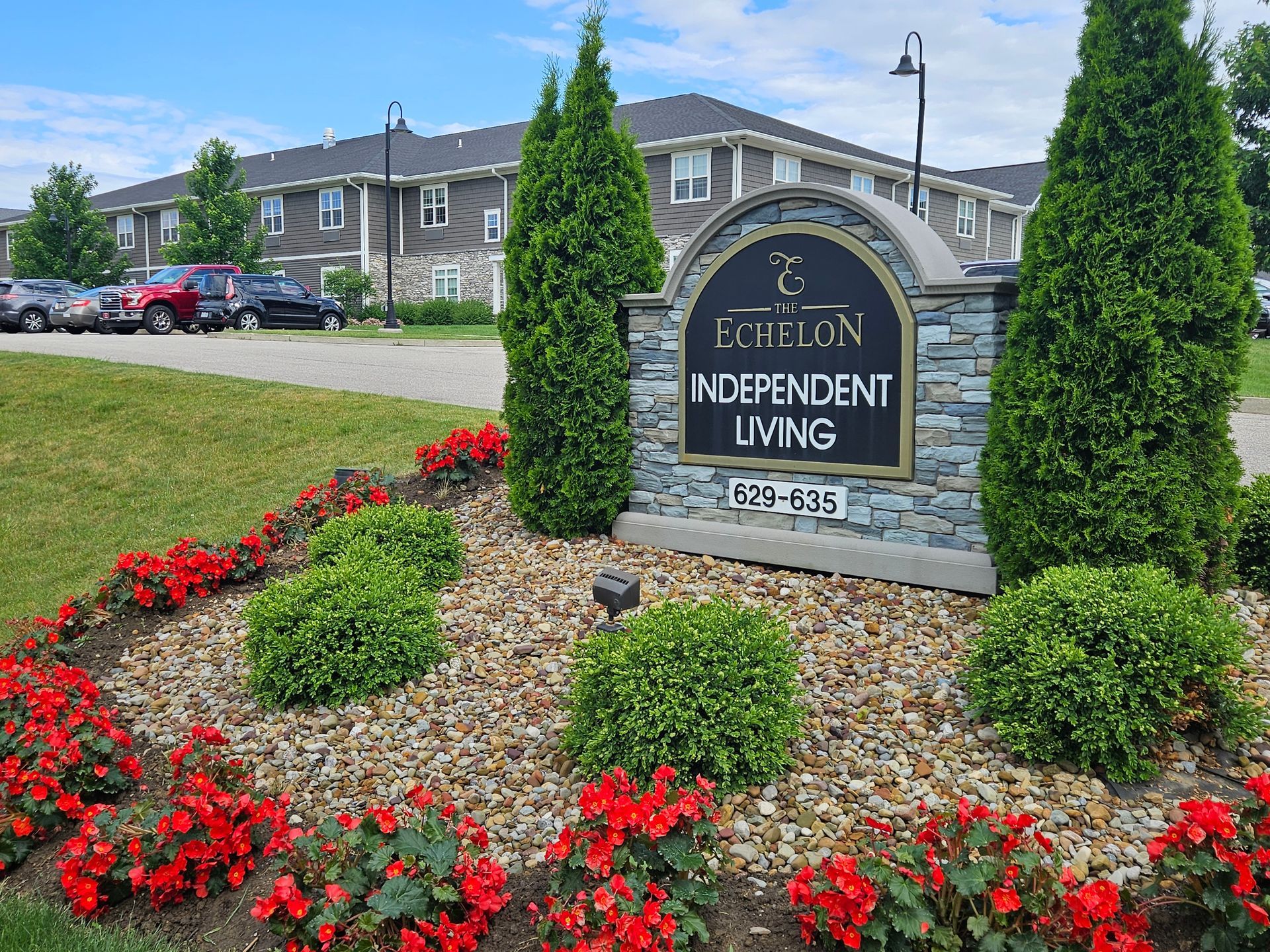 Sign for Exchelon Independent Living with building in the background, trees, and red flowers.