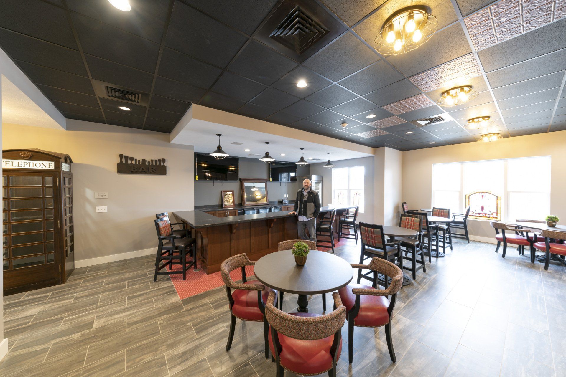 Lounge area with bar, tables, and chairs. Dark ceiling, gray floor. Phone booth on the left.