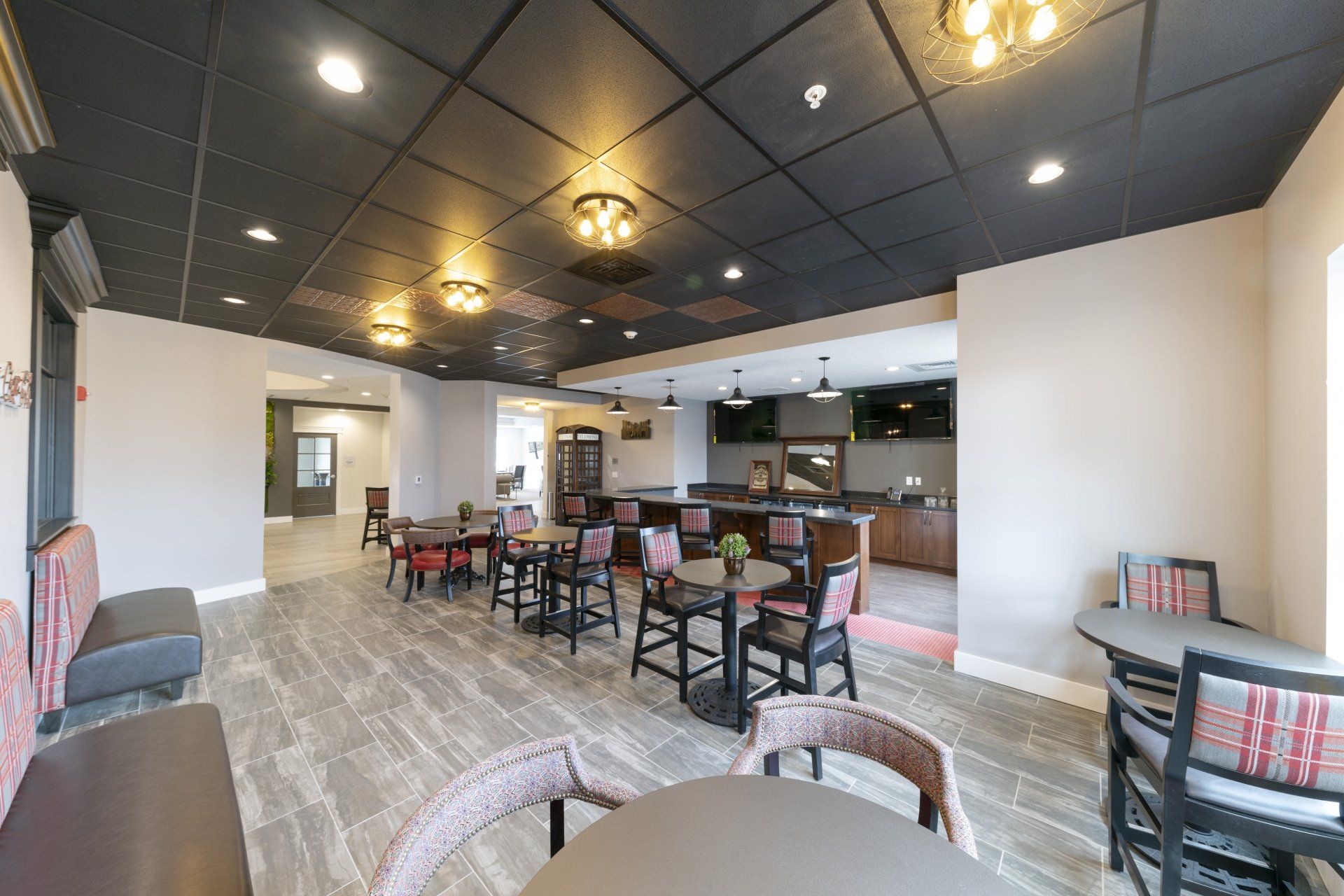 A dining area with tables, chairs, and a bar. Black ceiling, gray walls, and wooden flooring.