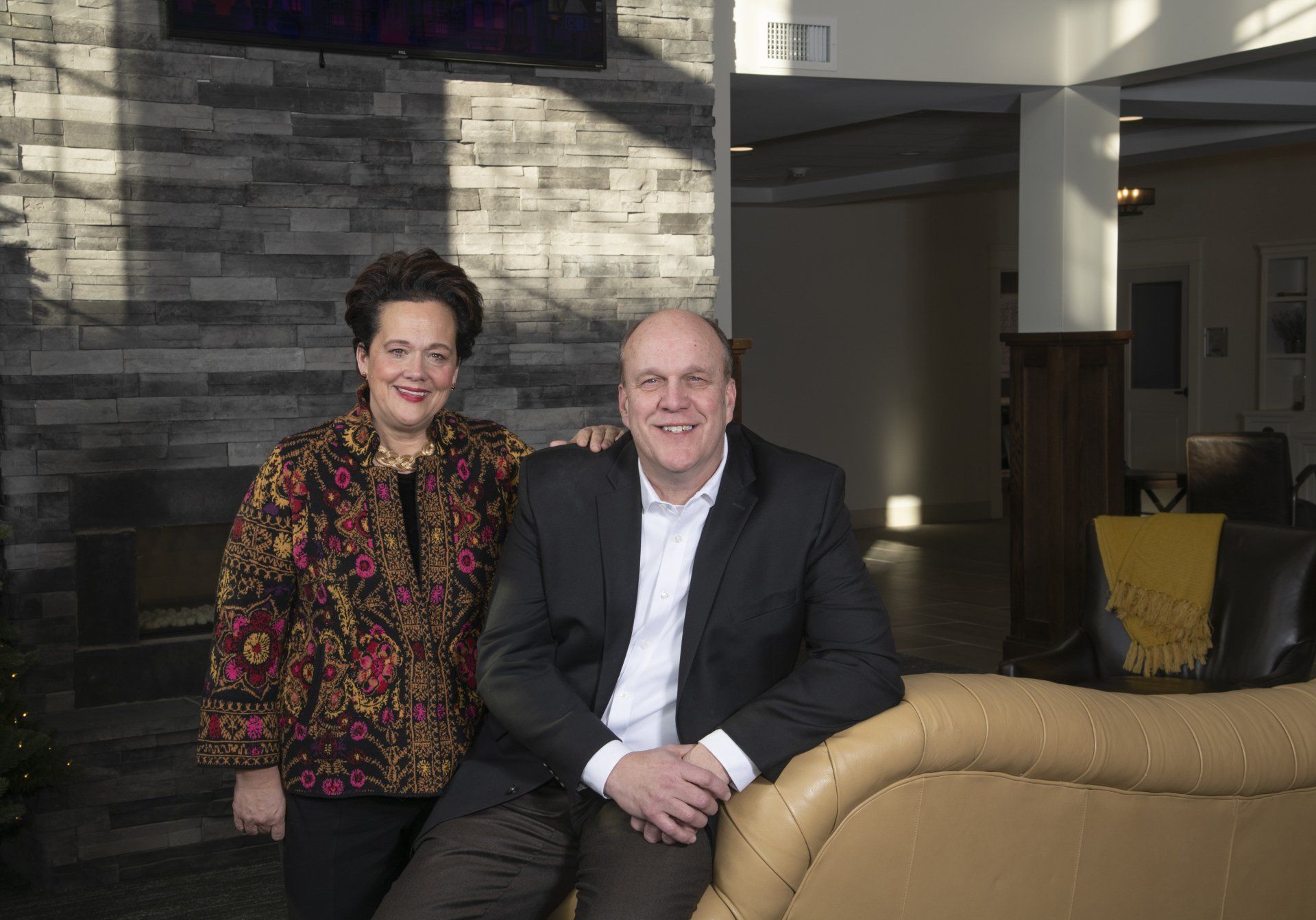 Woman in floral jacket and man in suit smiling together, leaning on a sofa. Interior setting.