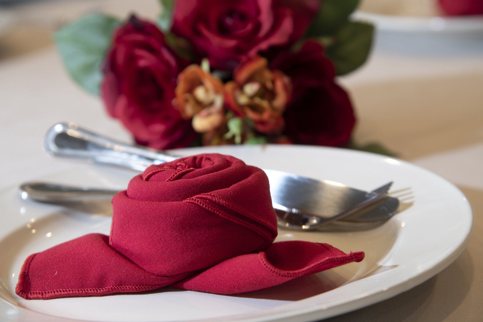 Red rose-shaped napkin on a white plate with silverware, and a red rose centerpiece.