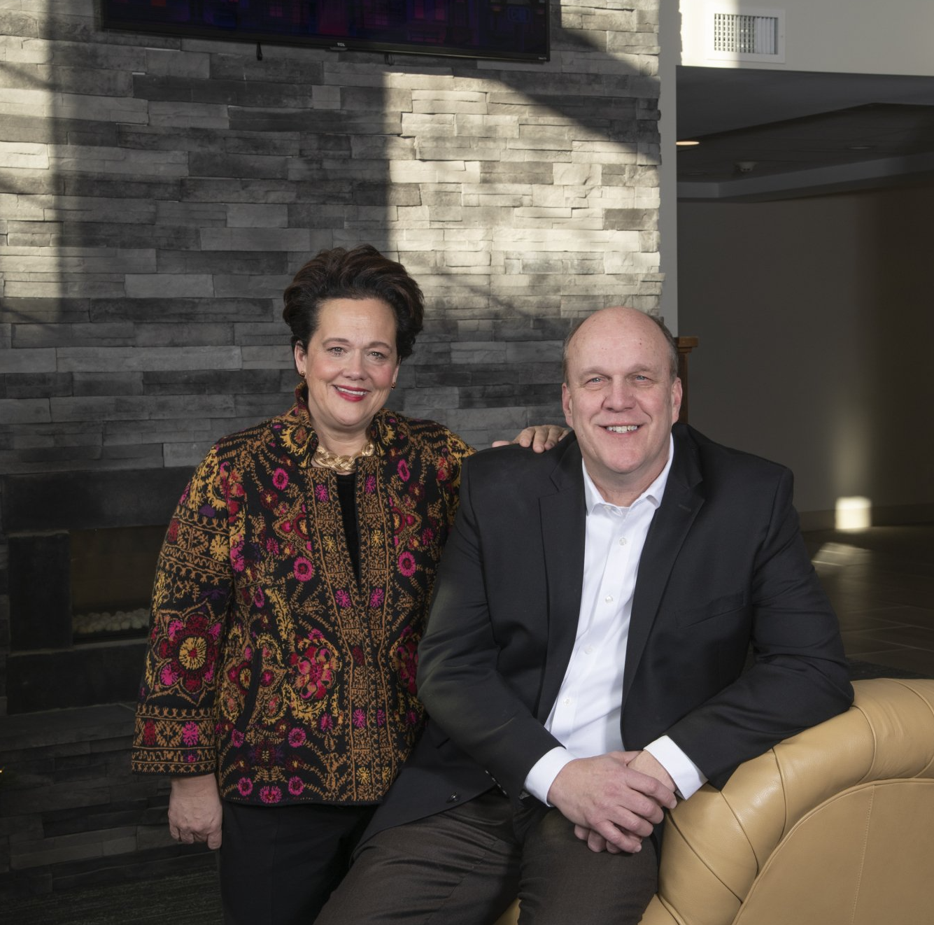 Woman in patterned jacket and man in suit smiling, posing indoors.