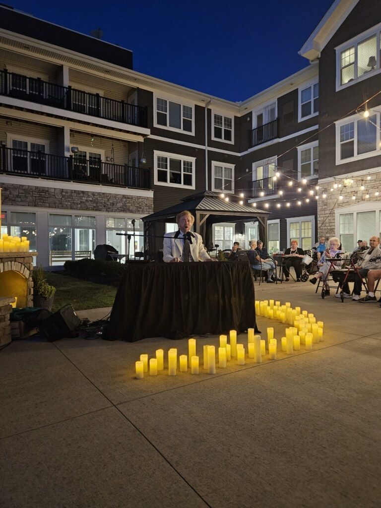 Person speaking at an outdoor event at night. Candles line the path, with a building in the background.