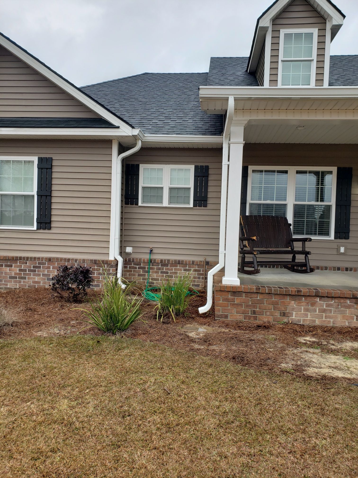 Tan house with dark shutters, white trim, and a porch, set in front of brown grass.