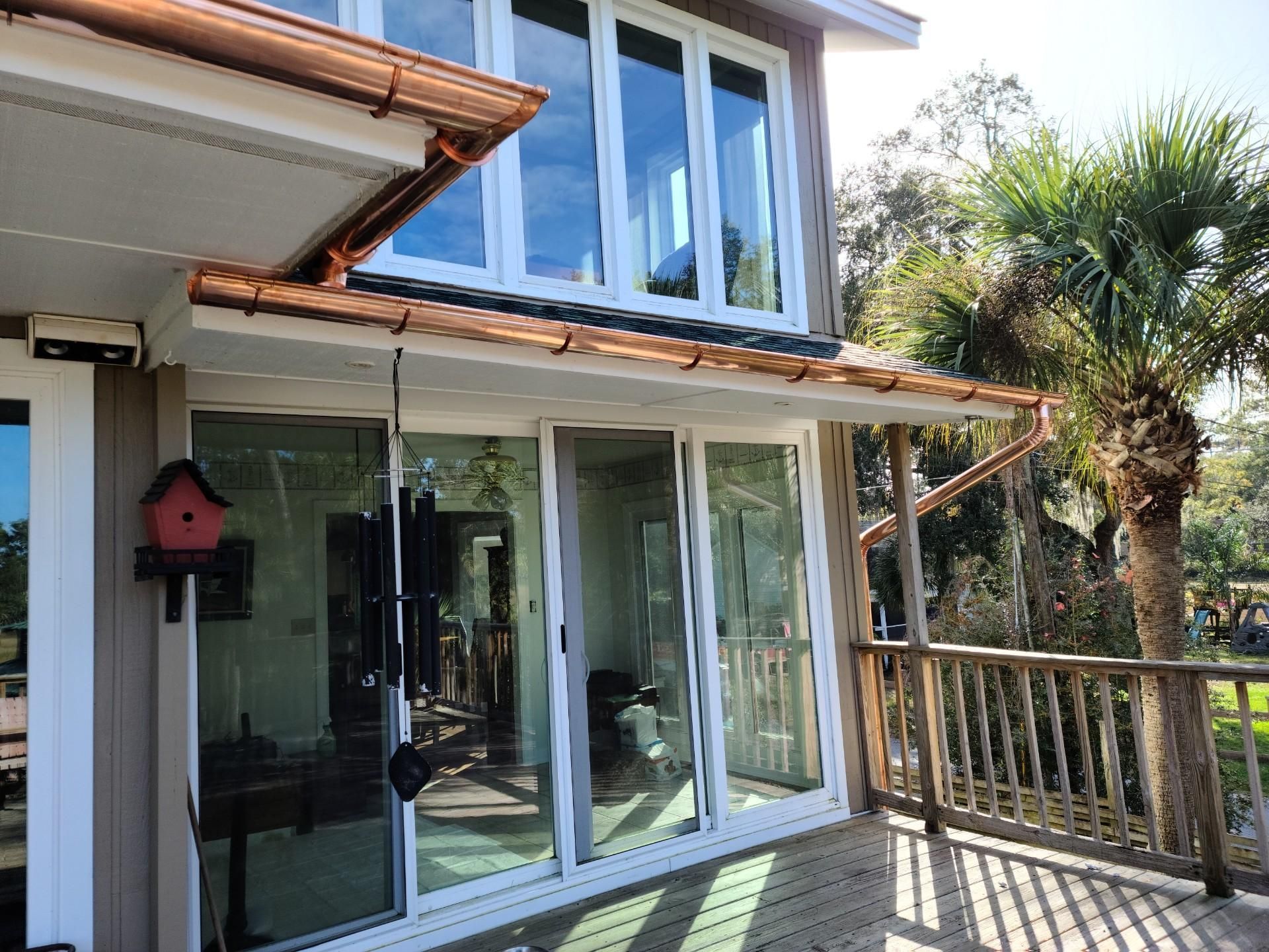 Two-story house with copper gutters, large windows, sliding doors, and a wooden deck. A palm tree is in the background.