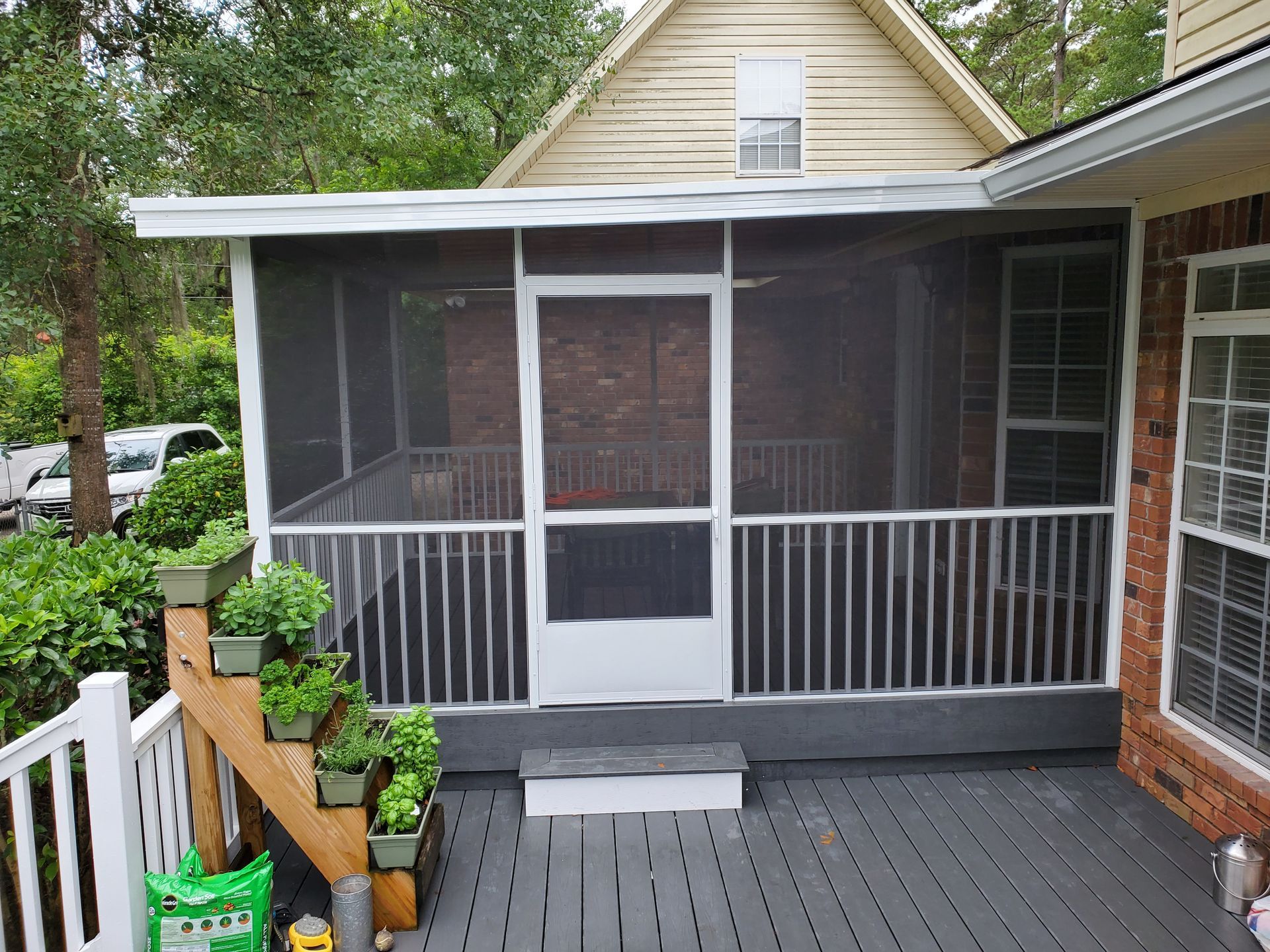 Screened-in porch with white railings, door, and trim. Dark gray deck and steps. Plants in pots on the deck.