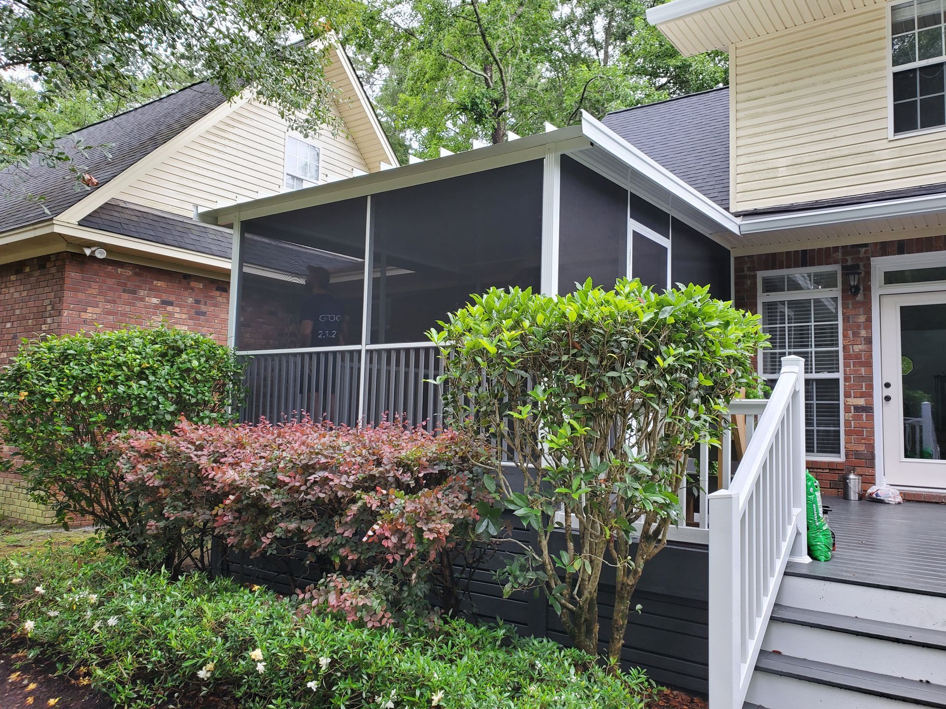 Screened porch attached to a house with white trim, dark screens, and surrounded by bushes and foliage.
