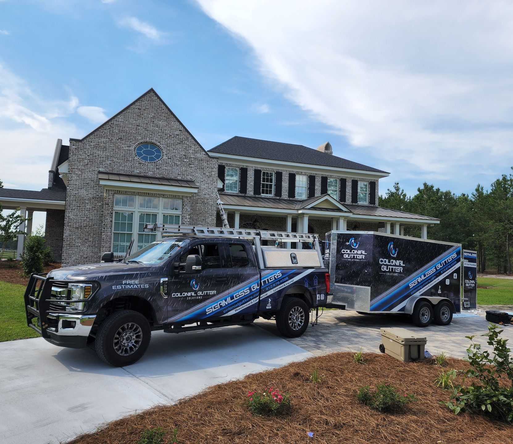 Truck and trailer with company logo parked in front of a large house with stone and siding facade. Sunny day.